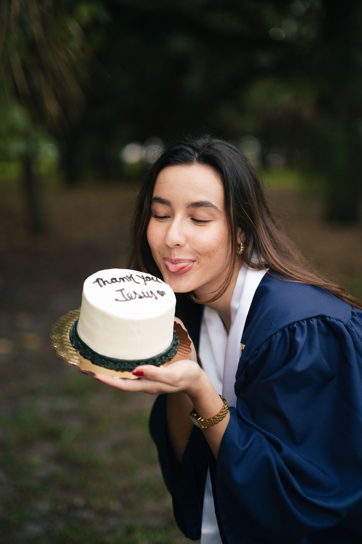Graduate in a navy gown playfully licks a white cake that reads thank you Jesus while standing in a wooded park.