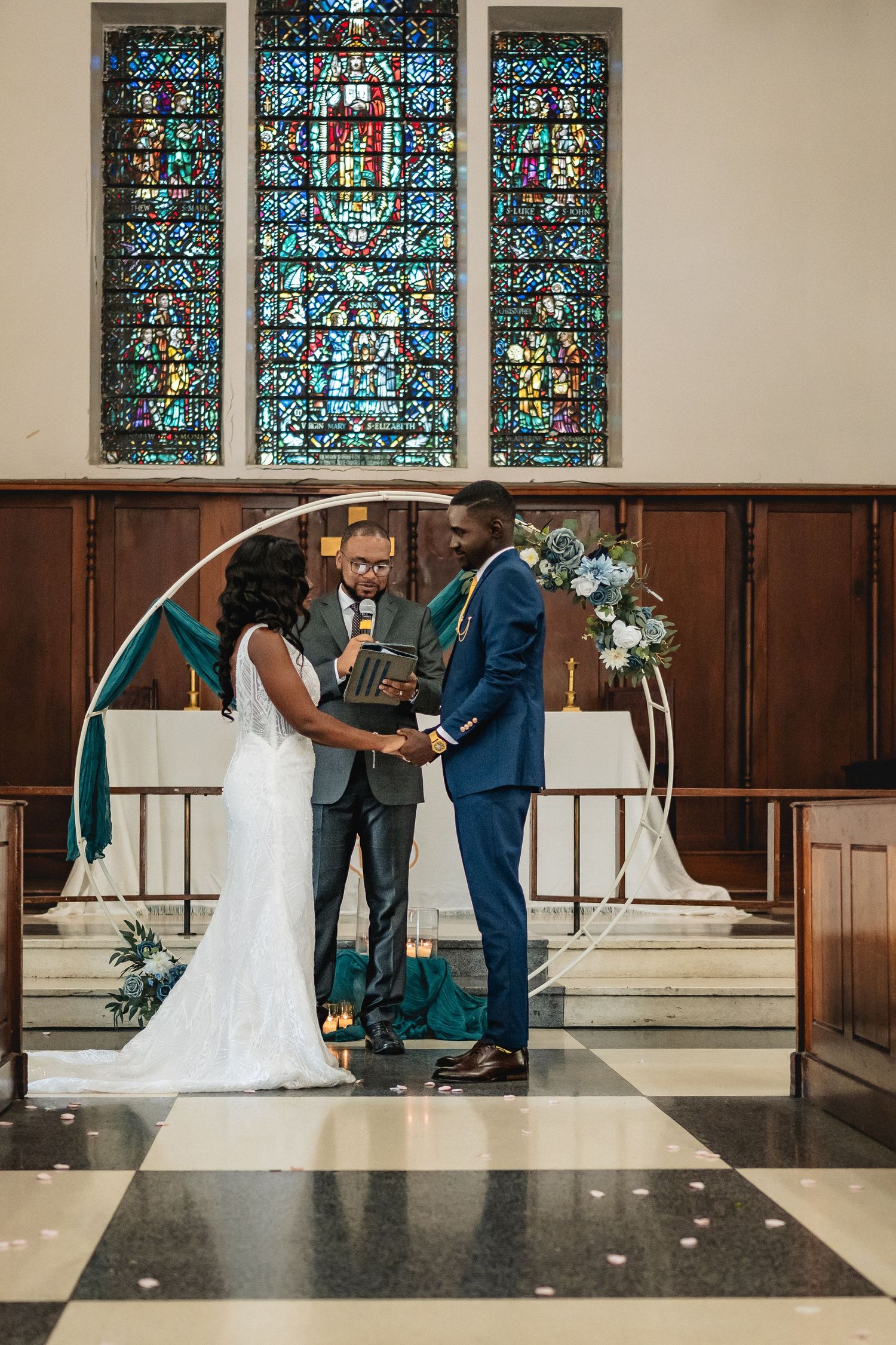 Couple exchanges vows at church altar with stained glass windows and circular floral arch.