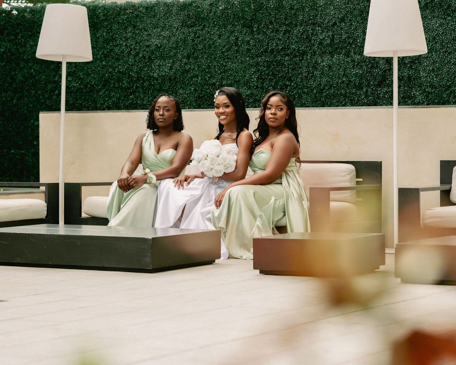 Bride in white gown seated with two bridesmaids in sage green dresses on an outdoor wedding patio.