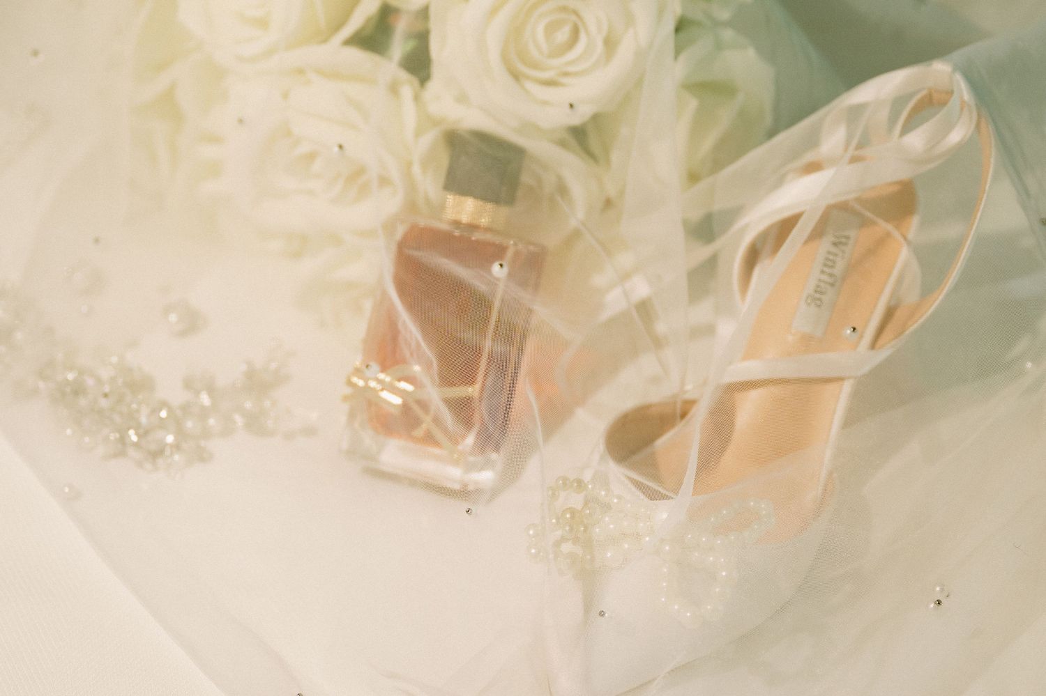 Elegant bridal accessories including white heels, perfume bottle, and pearl details on soft tulle veil.