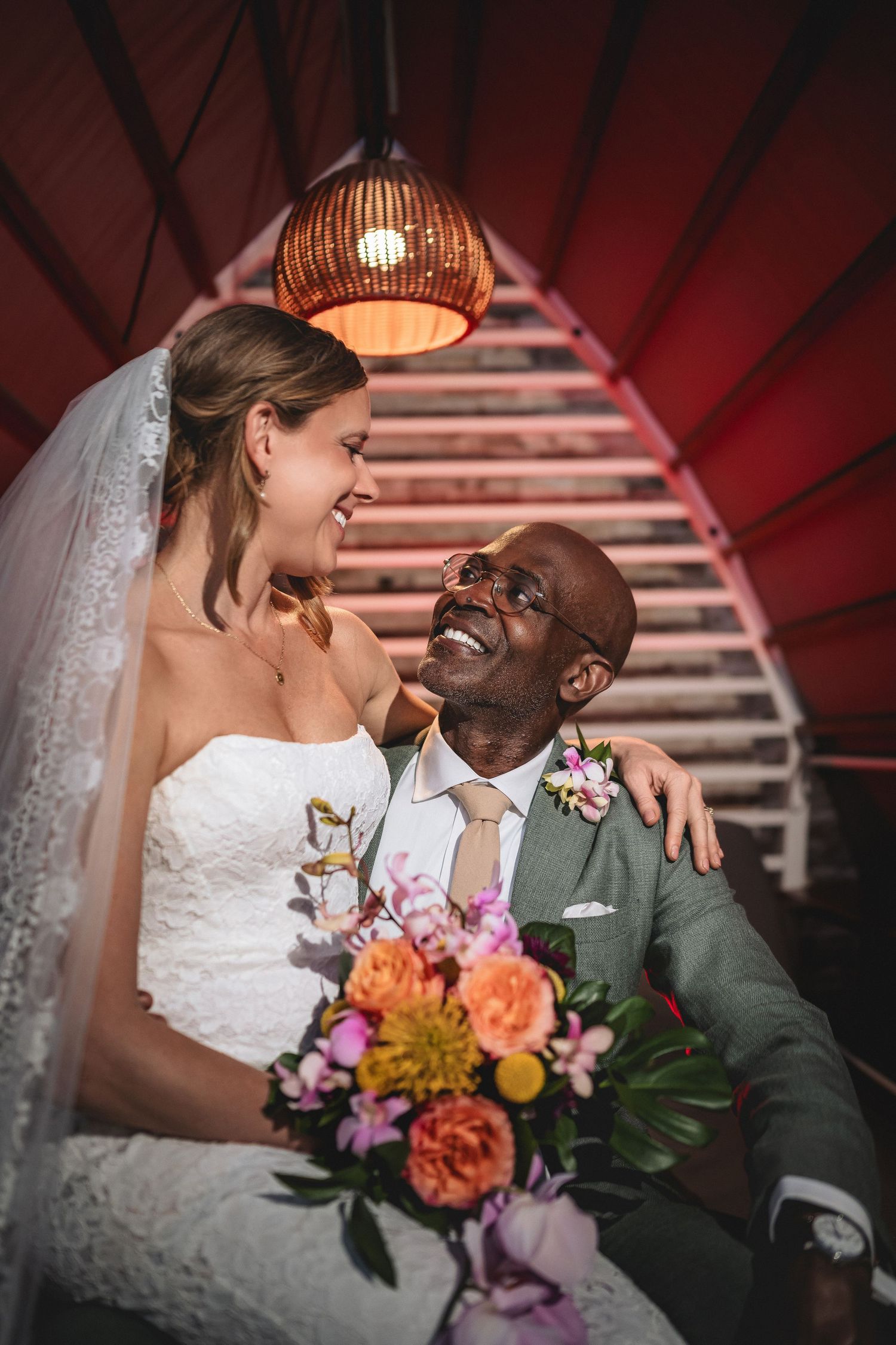 Joyful interracial couple in wedding attire sharing a loving moment under warm pendant lights with colorful bridal bouquet.