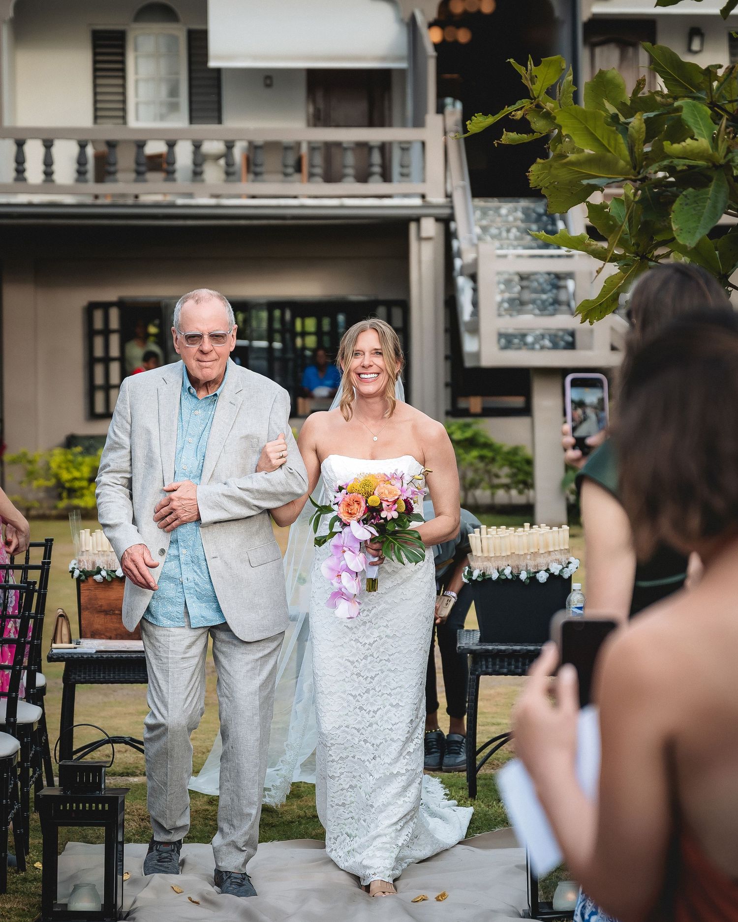 Bride in white gown holding colorful bouquet walks down aisle with older man in gray suit at outdoor wedding ceremony.