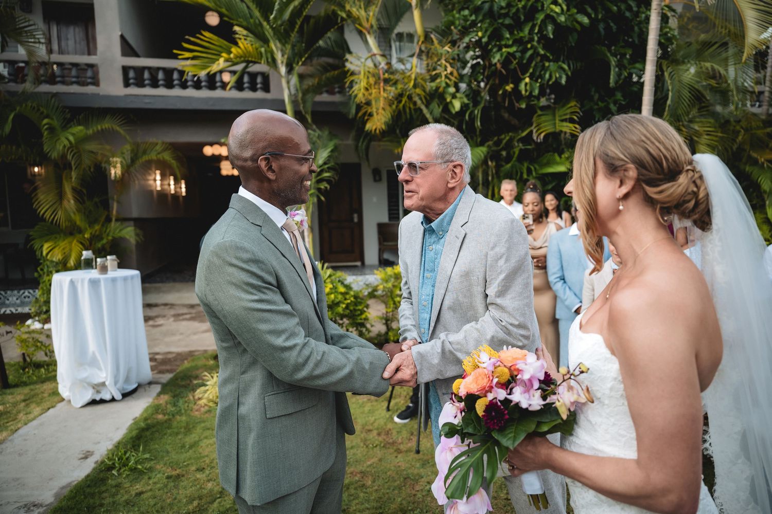 Bride holding colorful bouquet shakes hands with guest at outdoor tropical wedding ceremony.