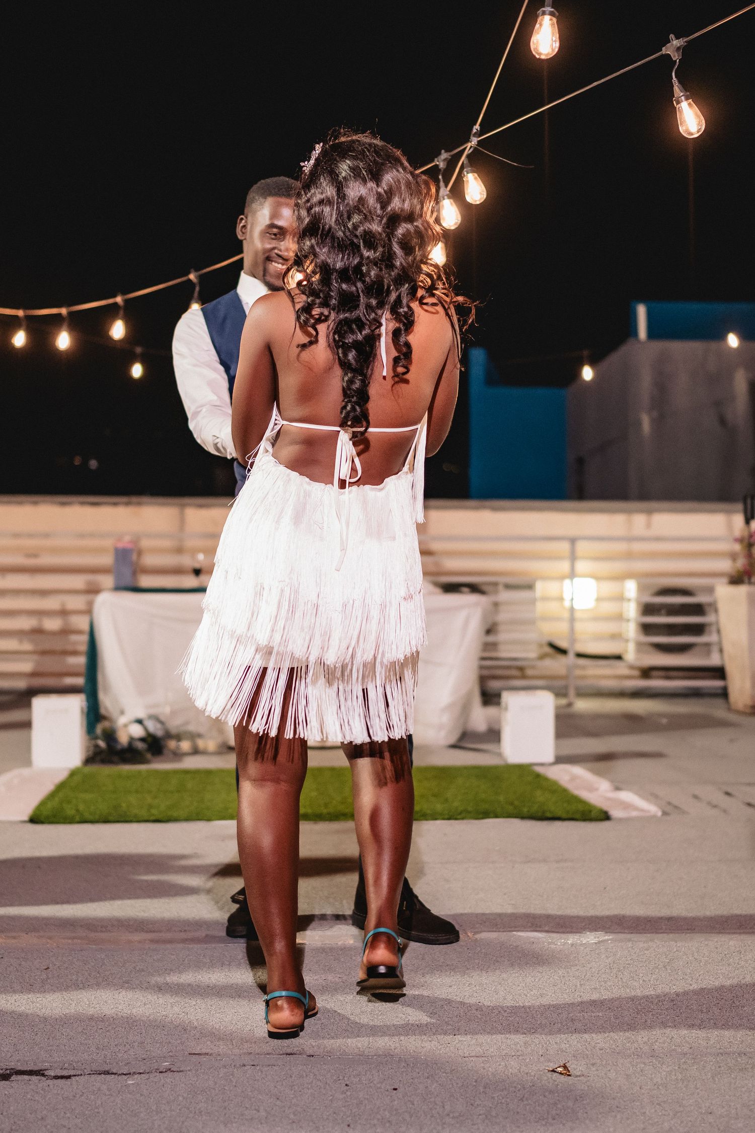 Bride in white fringe dress dances with groom under string lights at an outdoor evening wedding reception.