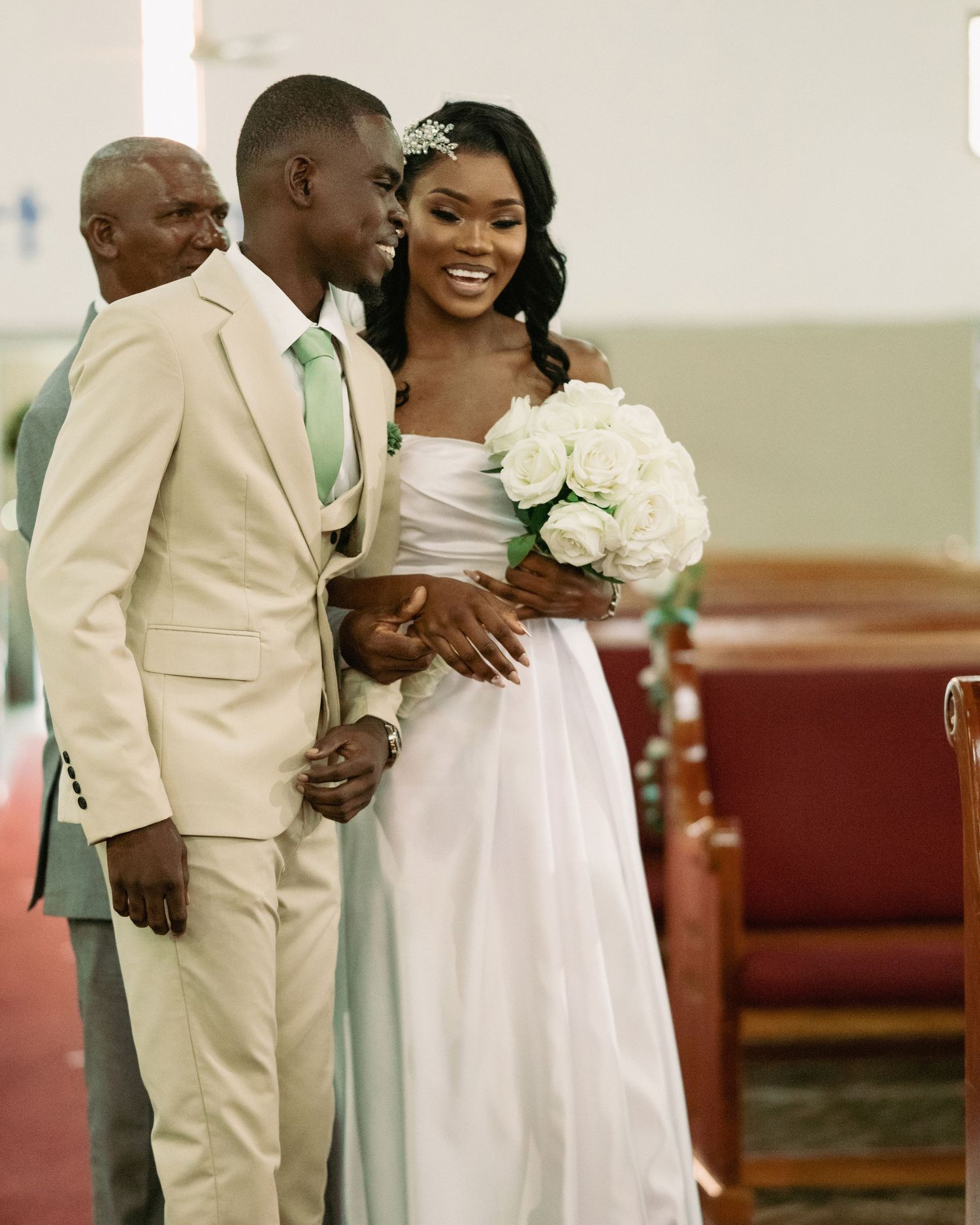 Smiling Black bride in white gown walks down church aisle with groom in beige suit and mint tie.