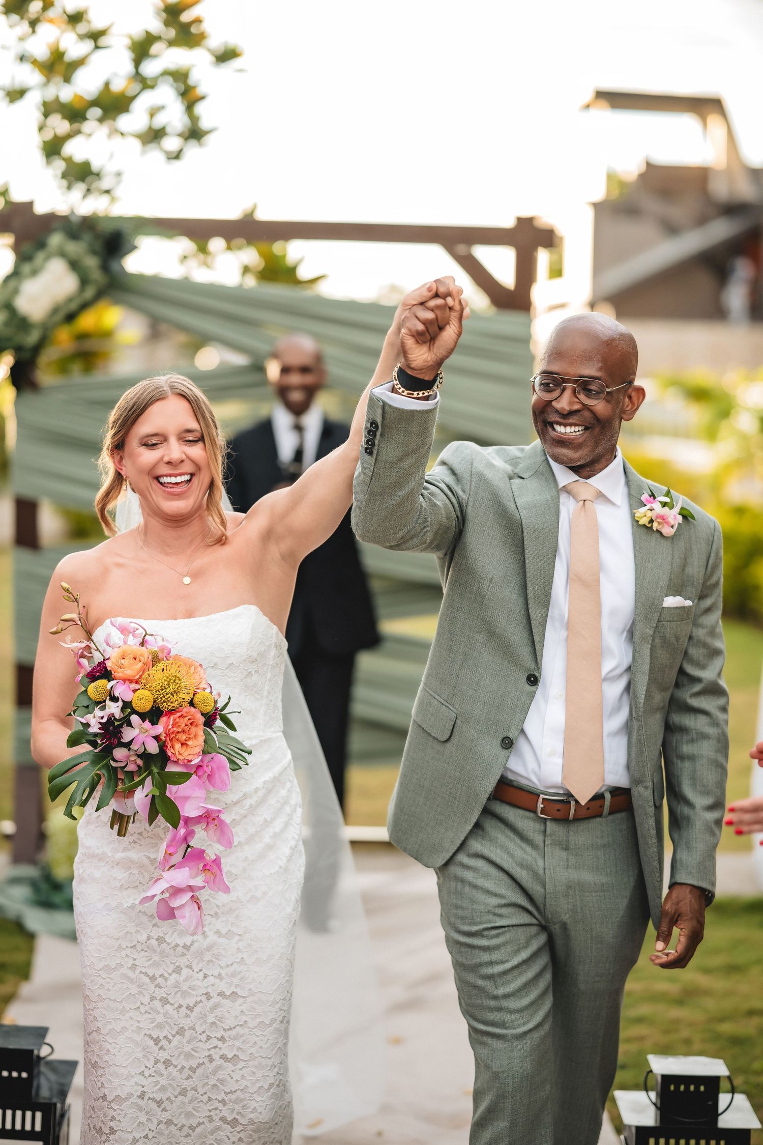 Joyful newlyweds celebrate after wedding ceremony, bride holding colorful bouquet, groom in sage suit raising hands.