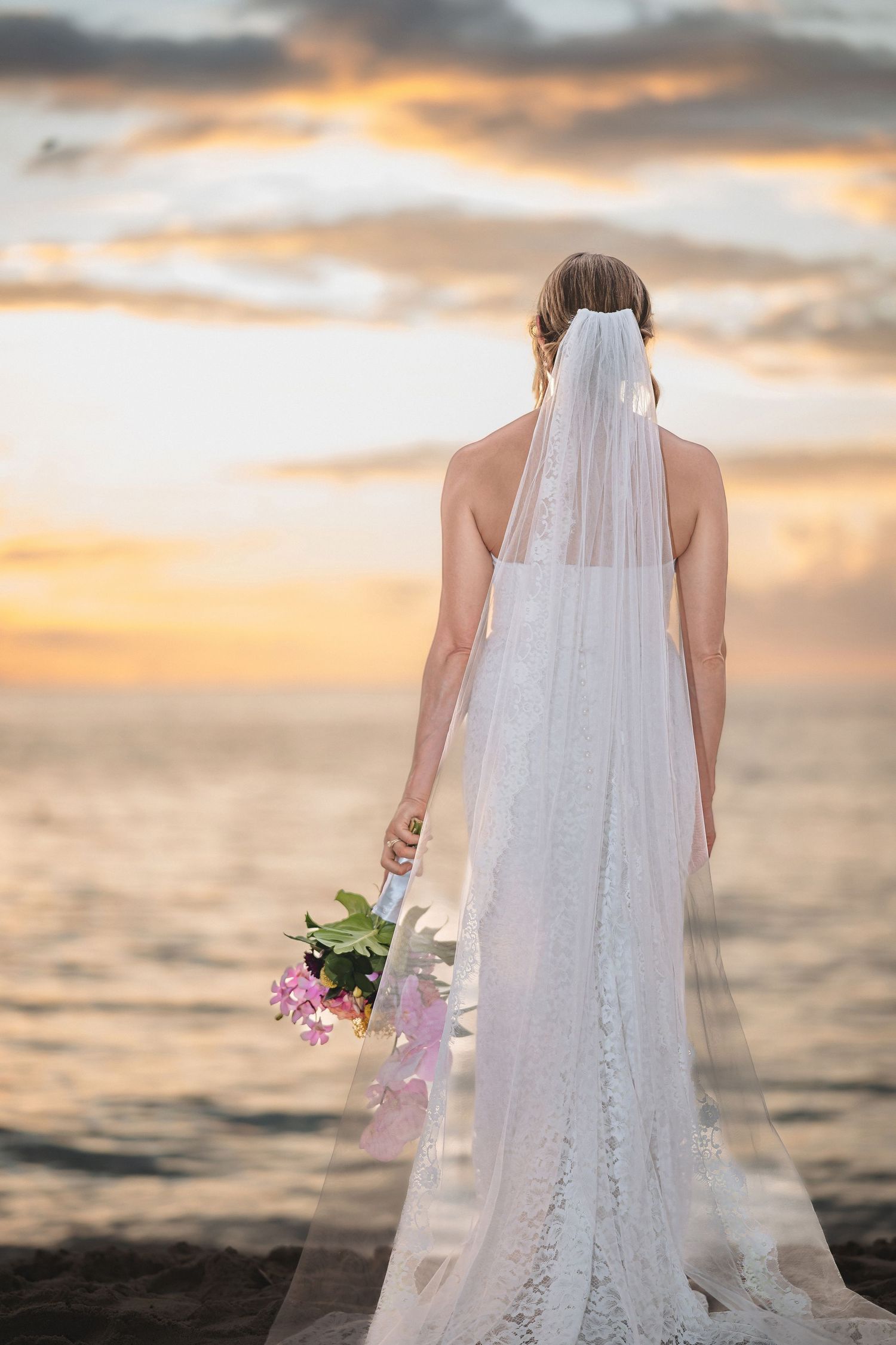 Bride in lace wedding dress and veil holding floral bouquet, standing by ocean at sunset.