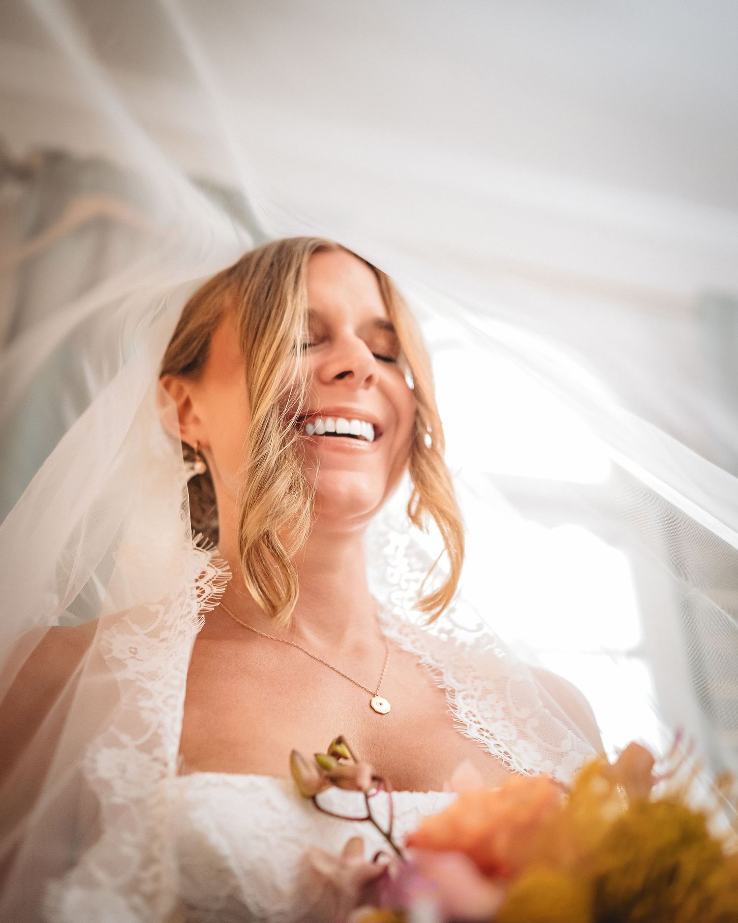 A radiant bride in a lace gown and veil laughs joyfully while holding a colorful bouquet on her wedding day.