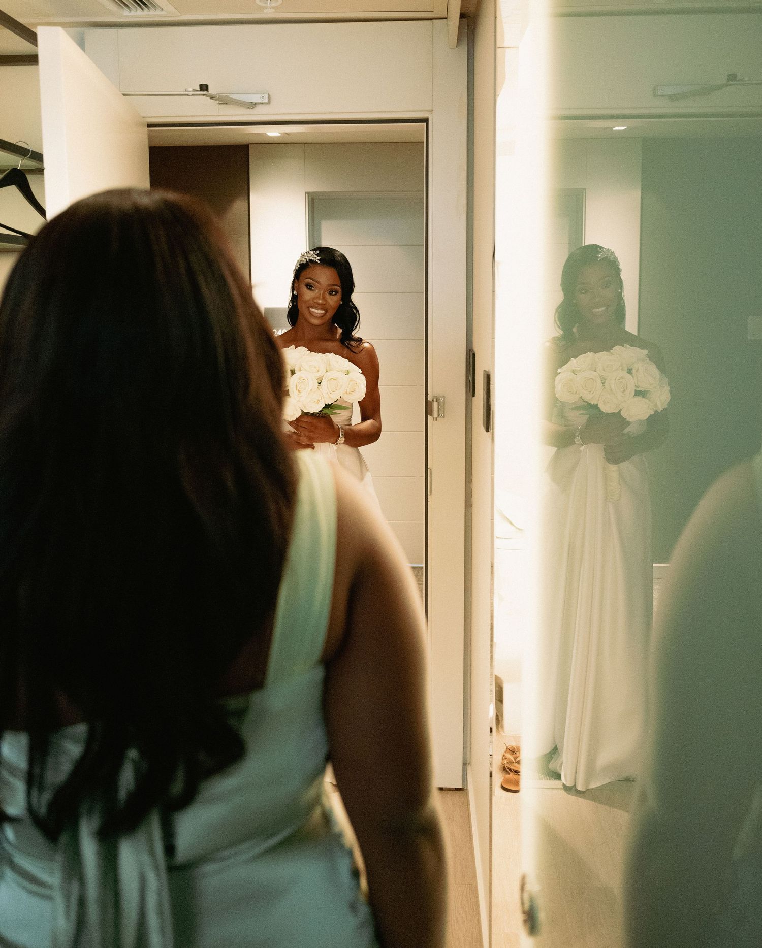 Radiant Black bride in white gown holding white bouquet reflected in mirror while bridesmaid looks on.