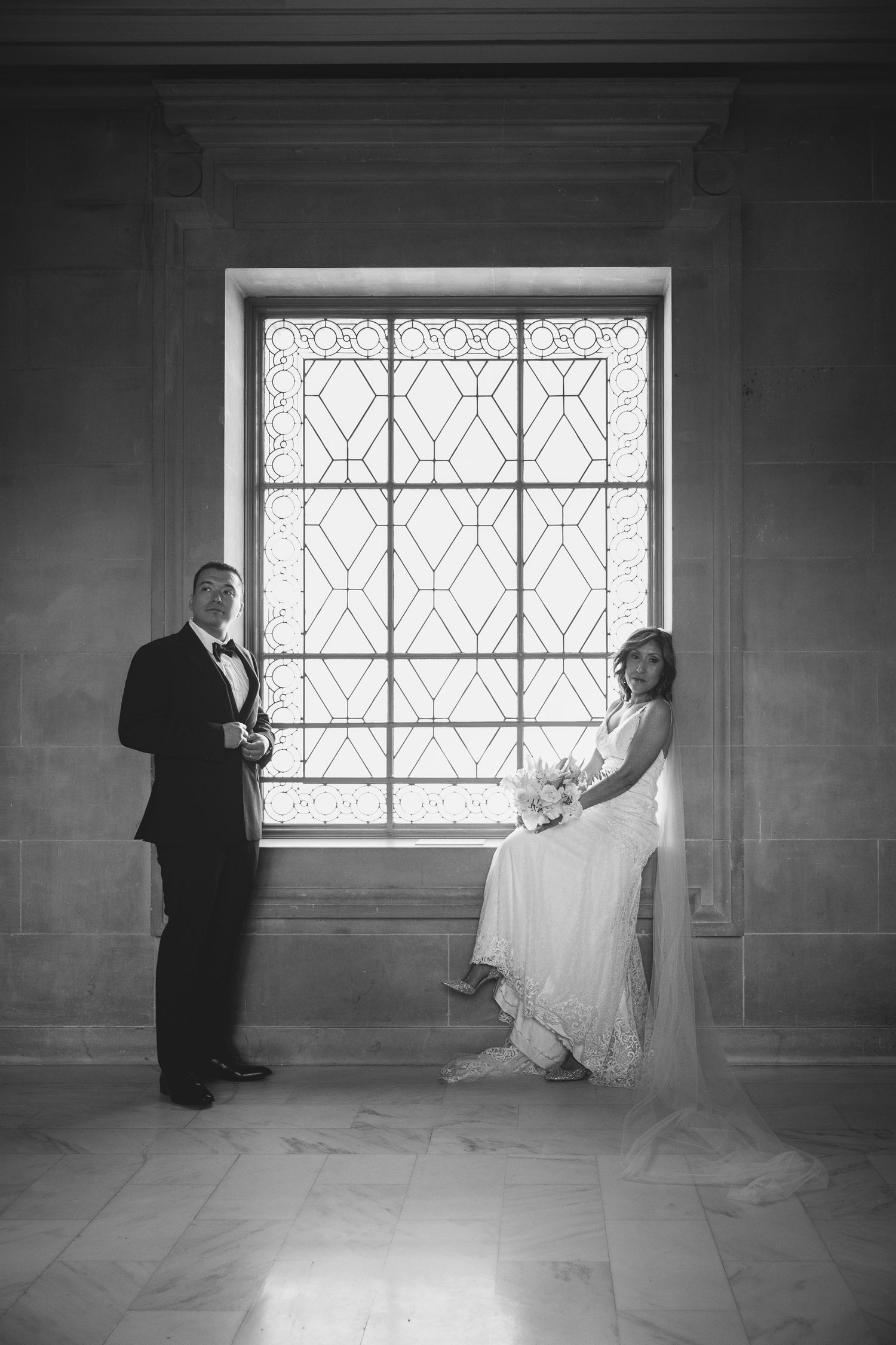 Black and white wedding portrait of bride and groom standing by ornate stained glass window in elegant venue.
