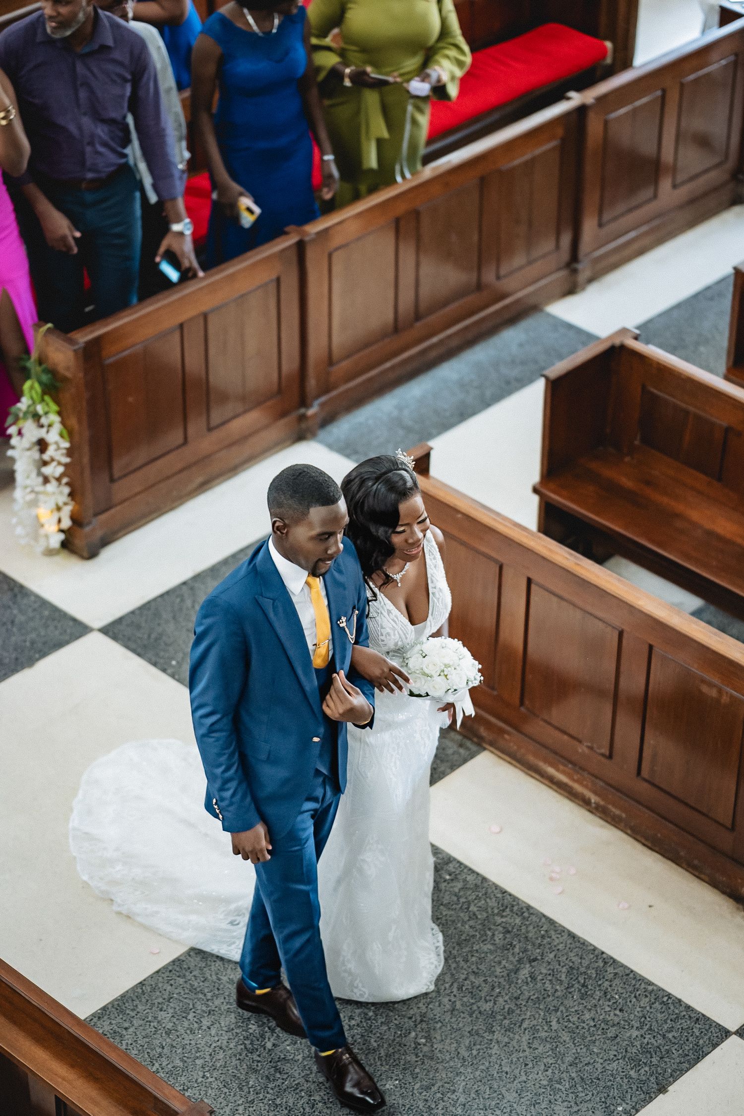 Bride and groom walk down the aisle of a church, the groom in a blue suit and gold tie, bride in a white gown.