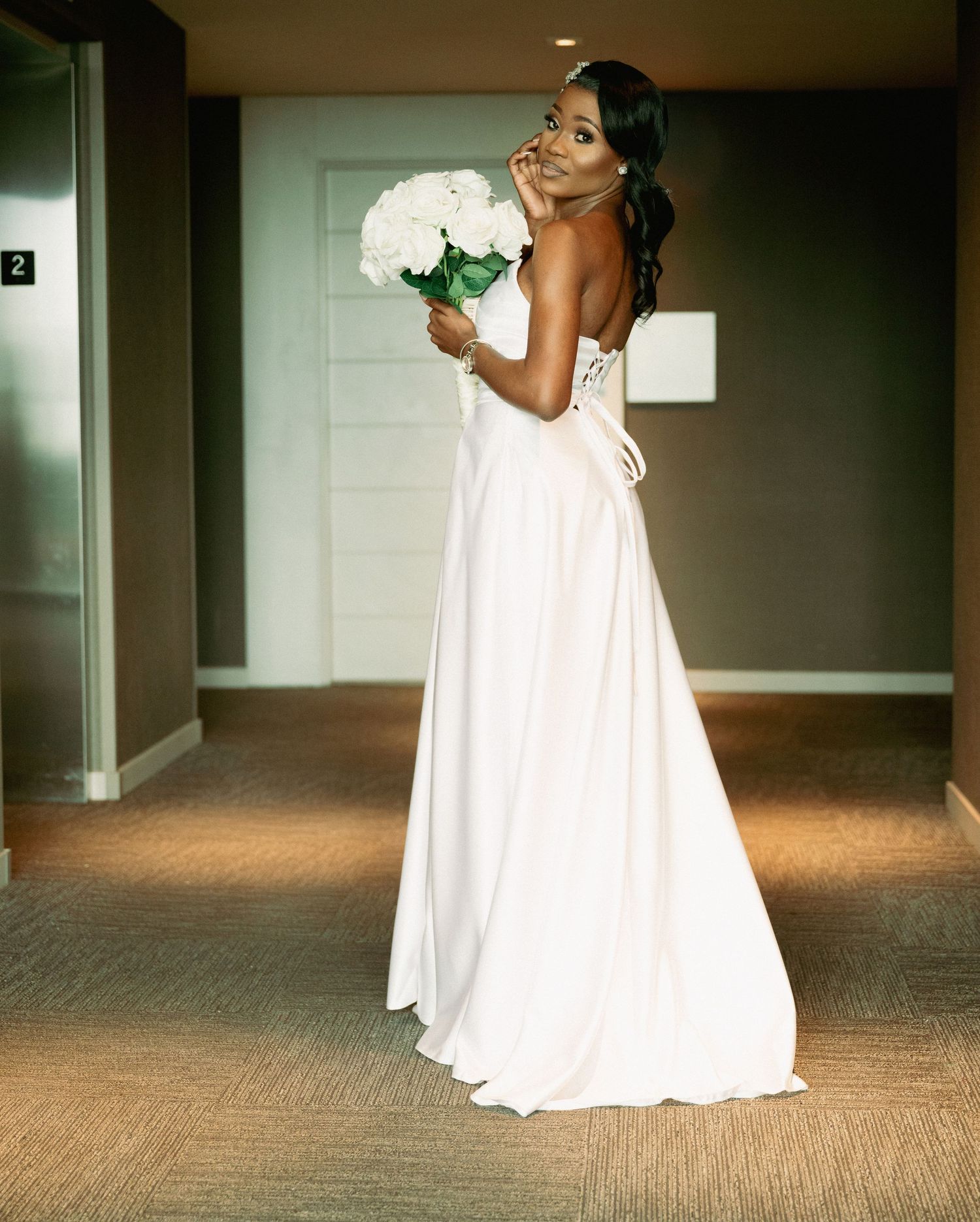 A bride in a white gown holds a white bouquet while posing in a hallway.