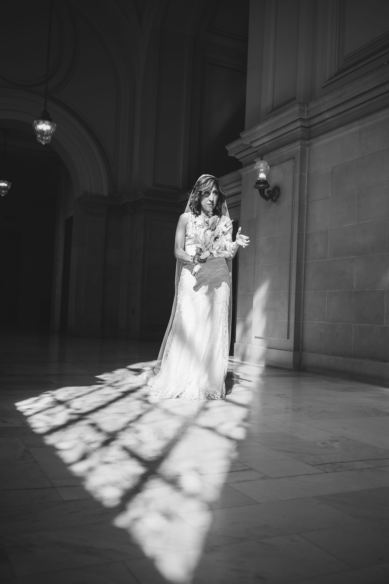 Black and white photo of a bride in a flowing gown and veil standing in sunlight inside an elegant hall.