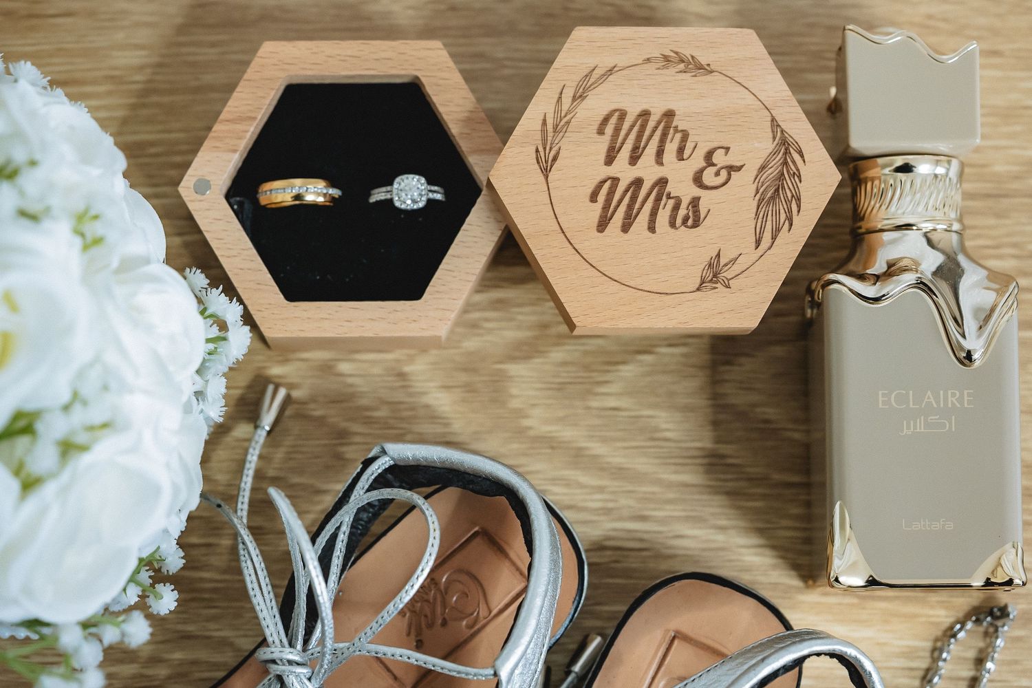 Hexagonal wooden Mr & Mrs ring box with wedding bands, perfume bottle, and silver sandals on wooden surface.