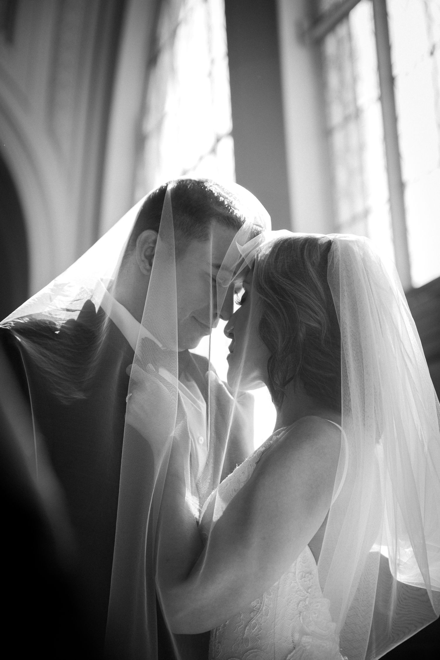 Black and white wedding photo of bride and groom sharing intimate moment under veil in church.
