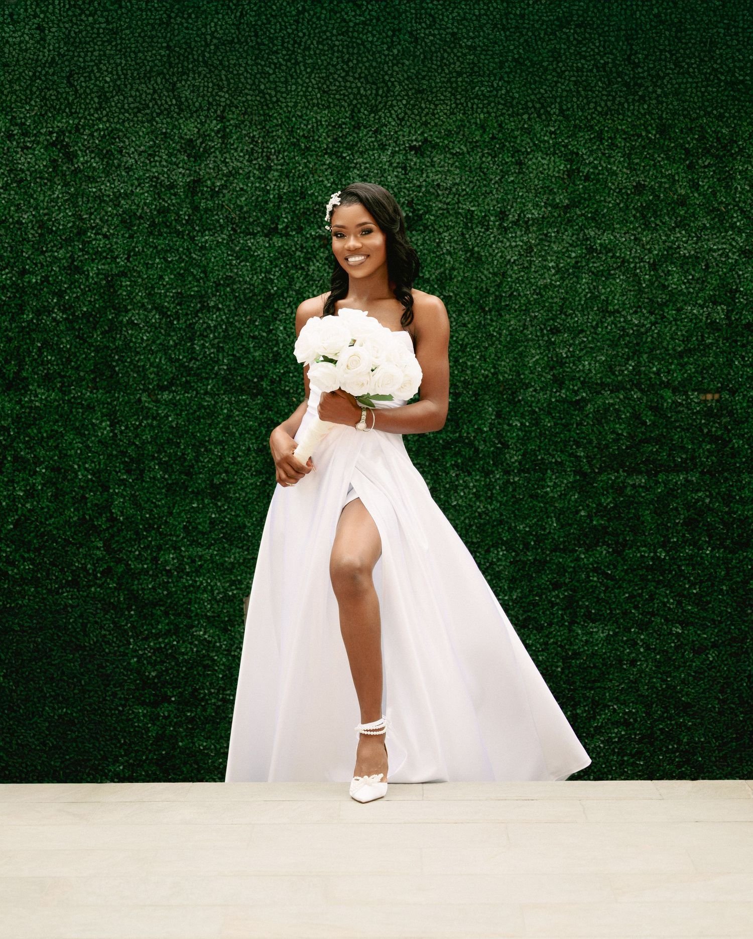 A smiling bride in a white gown holds a bouquet in front of a green hedge wall.