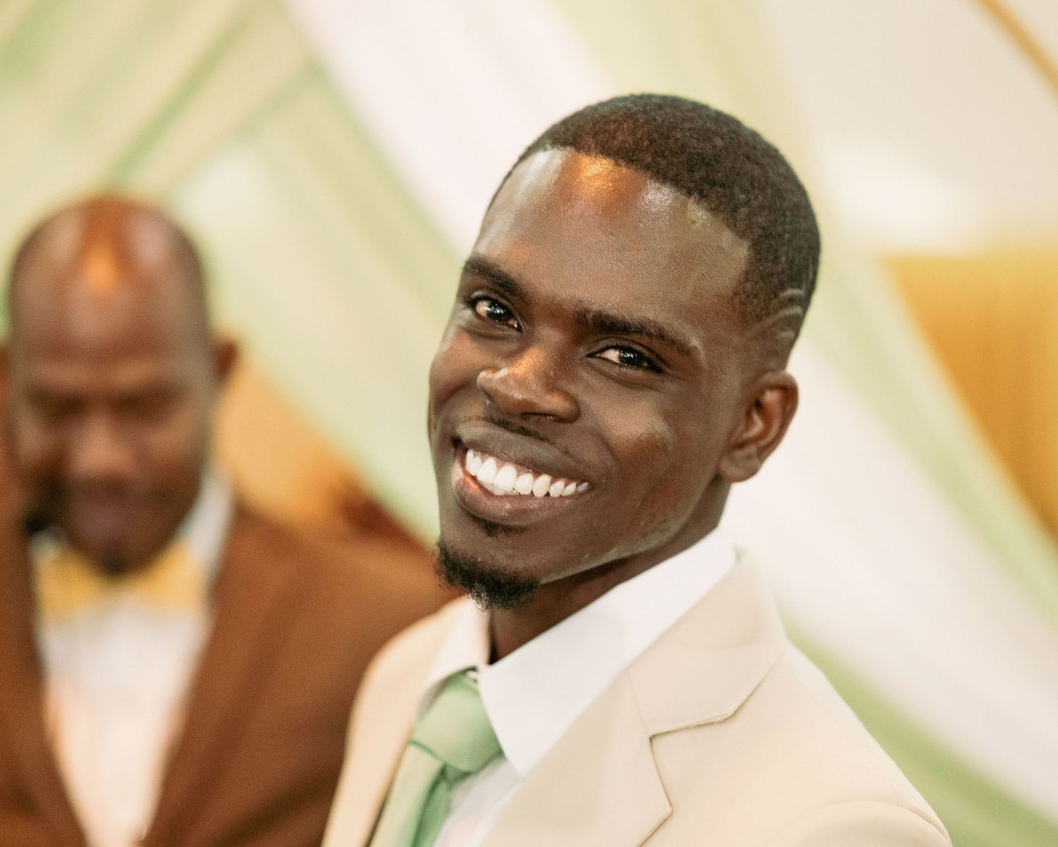 Smiling groom in beige suit with green tie at outdoor wedding celebration.
