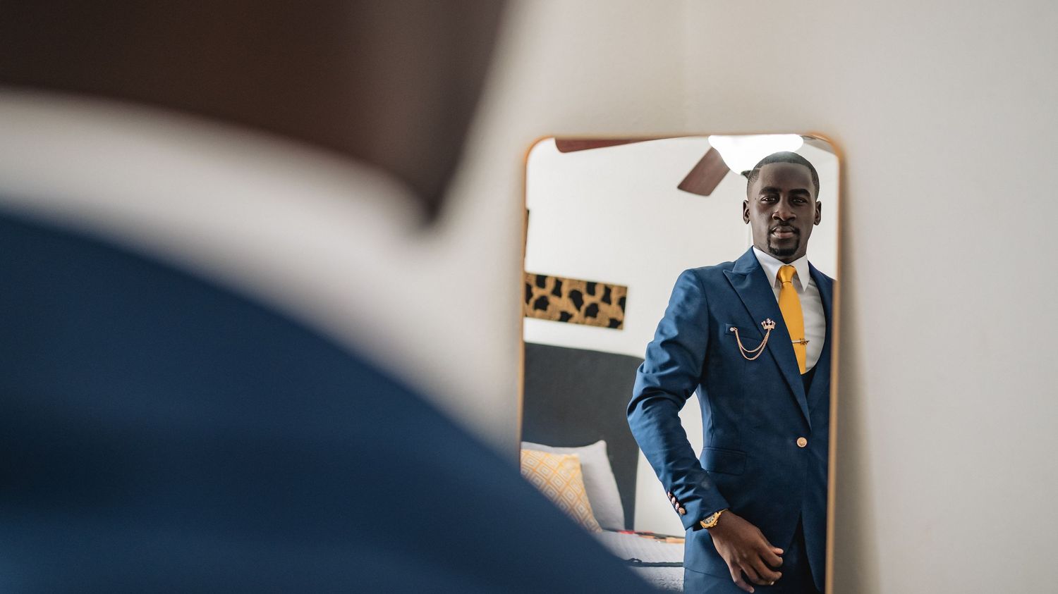 Groom in navy blue suit with yellow tie checks his appearance in a mirror before his wedding ceremony.