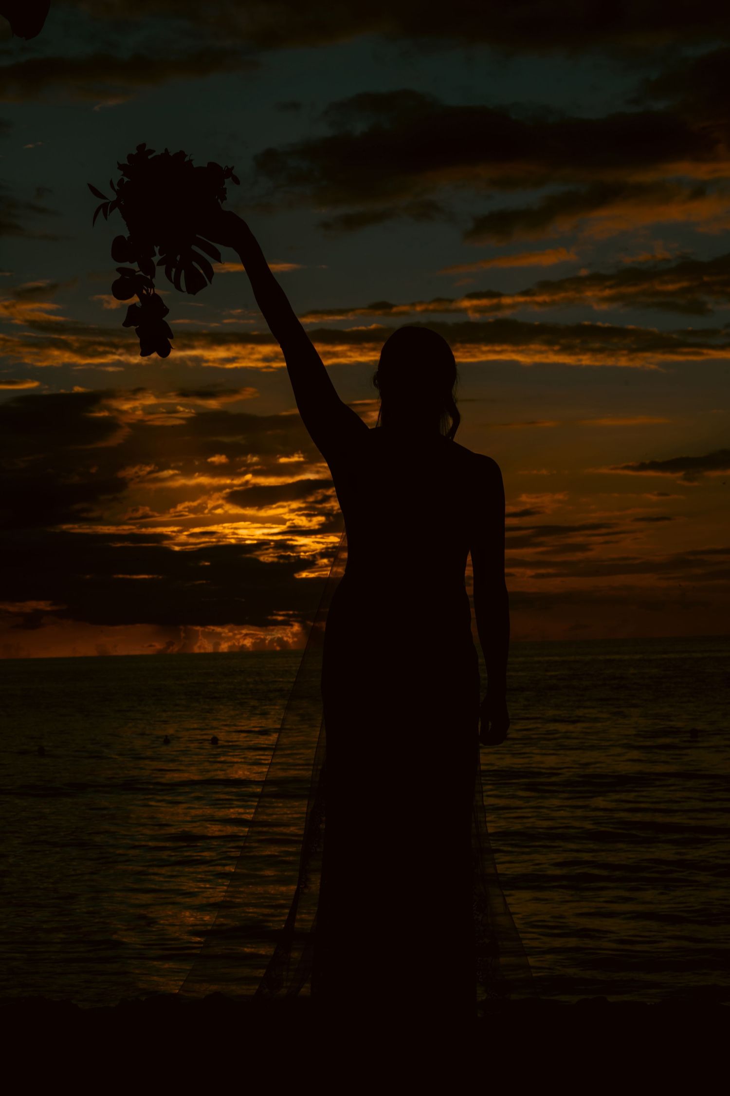 Silhouette of a bride holding a bouquet aloft against a dramatic ocean sunset with glowing orange clouds.
