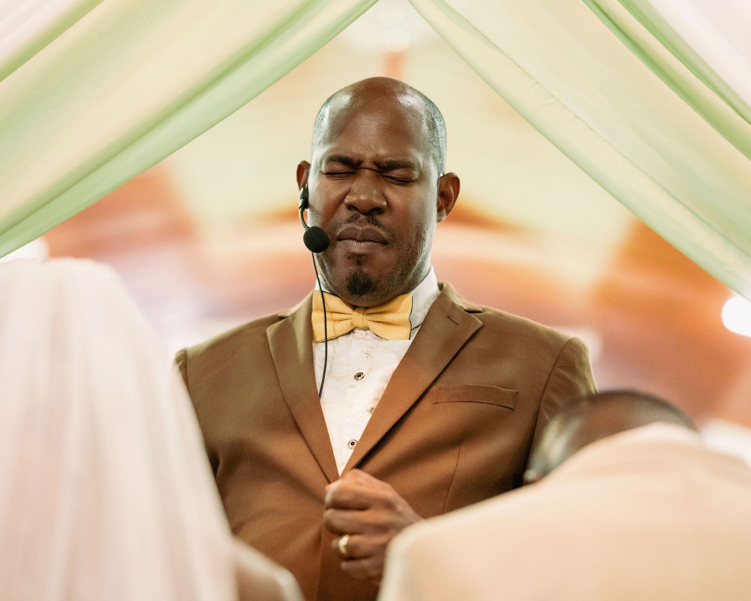 Man in brown suit with yellow bow tie and headset microphone officiates a wedding ceremony indoors.