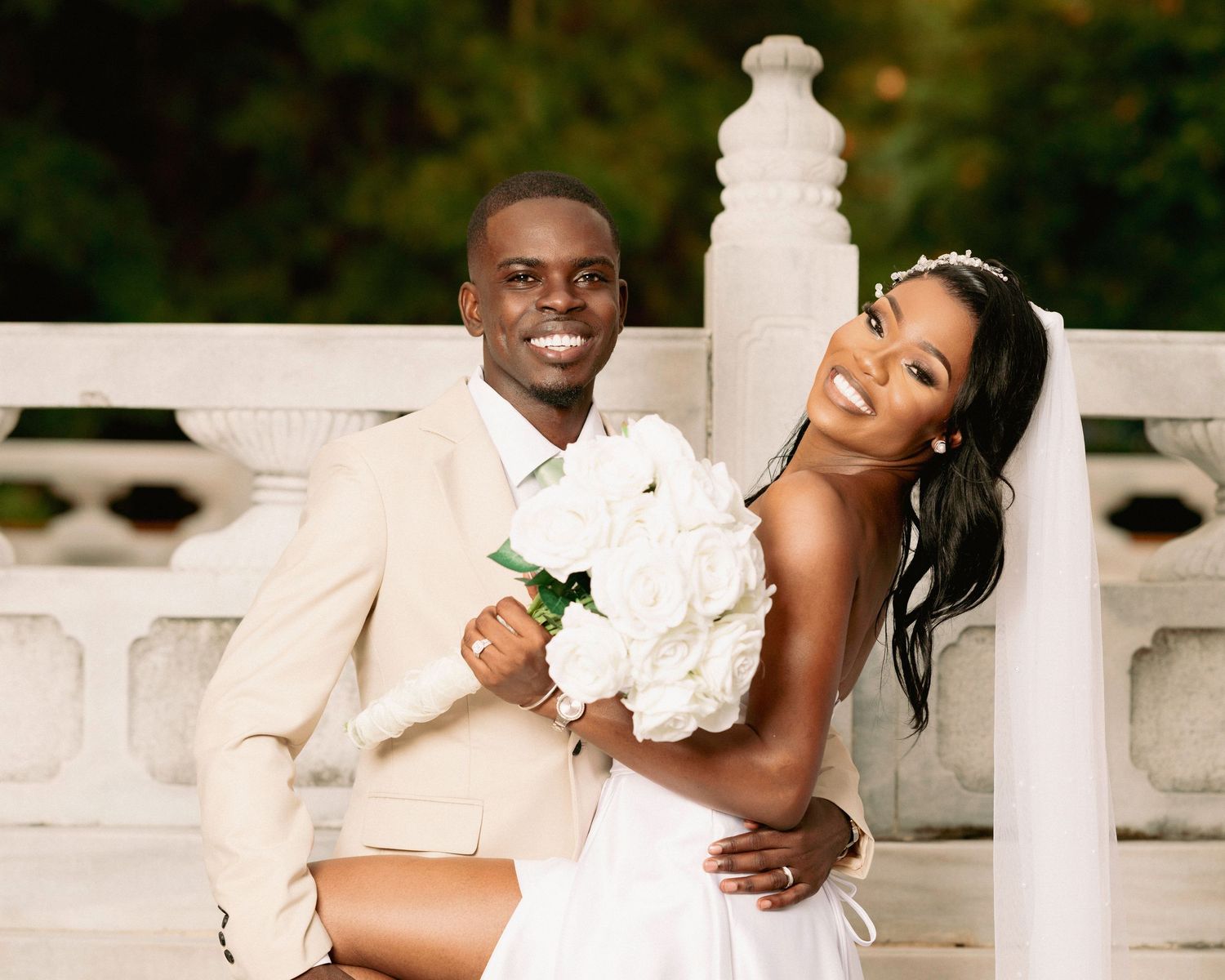 Happy newlyweds pose together, groom in beige suit holding bride in white gown with bouquet of white roses outdoors.