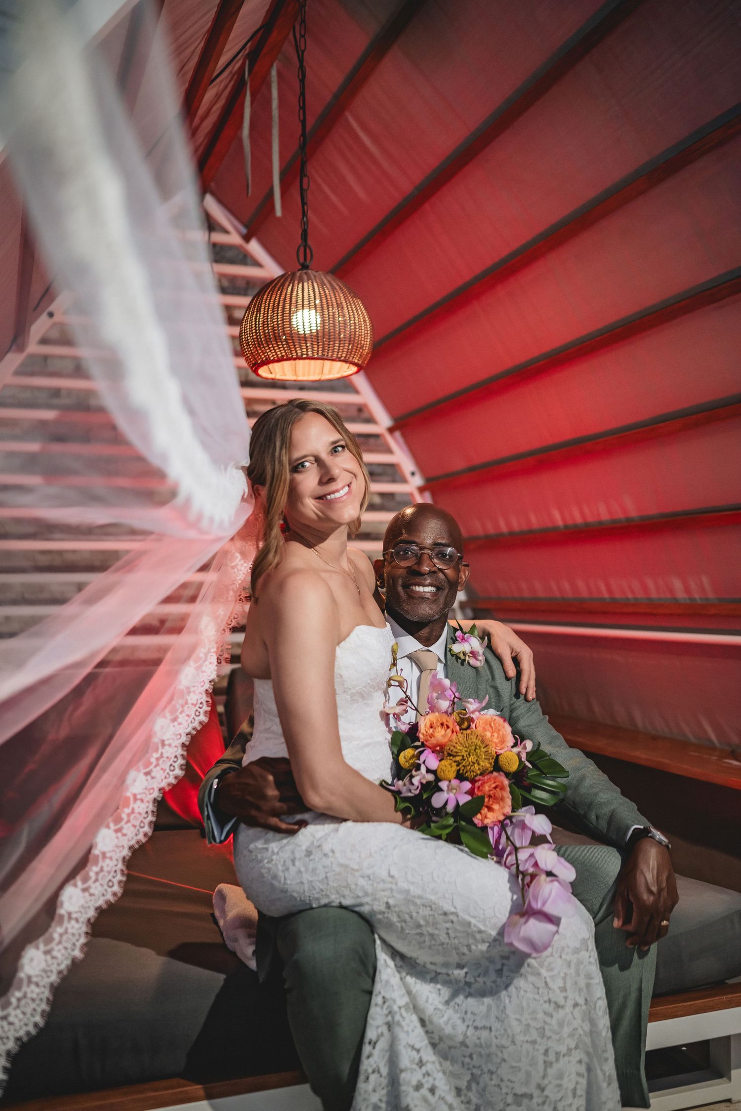 Interracial couple in wedding attire sitting together, bride in white lace gown holding colorful bouquet, groom in gray suit.