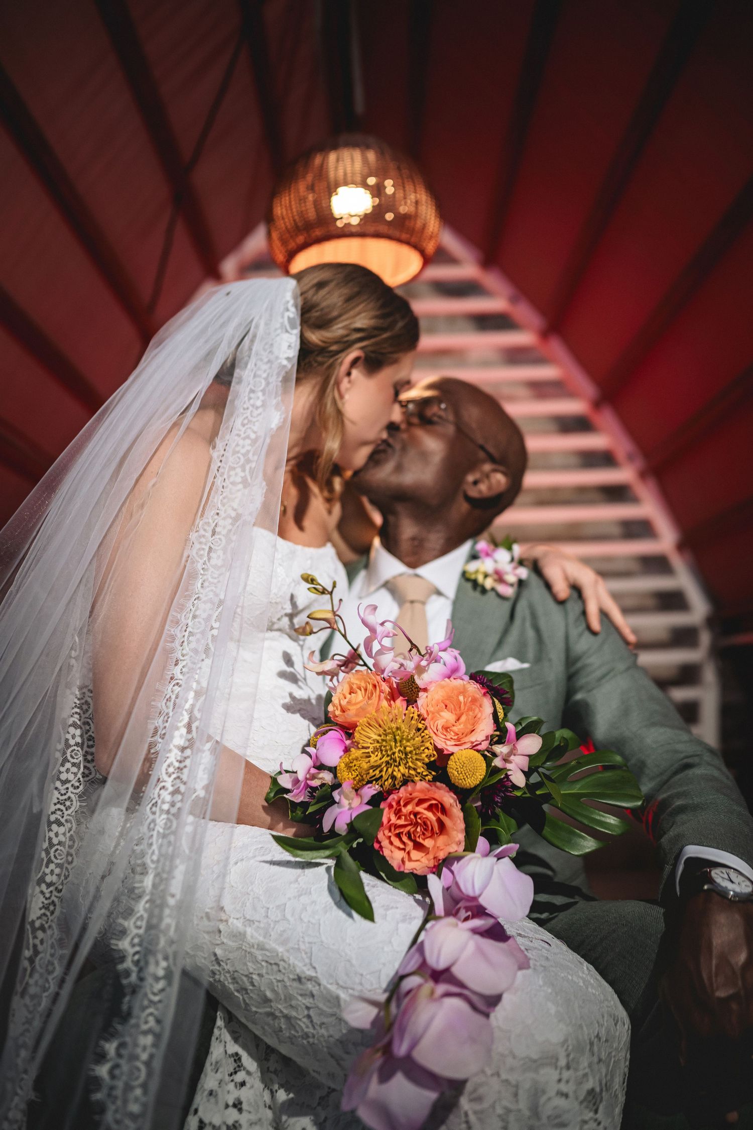 Interracial couple sharing a kiss on their wedding day, bride in lace gown with veil holding colorful bouquet.