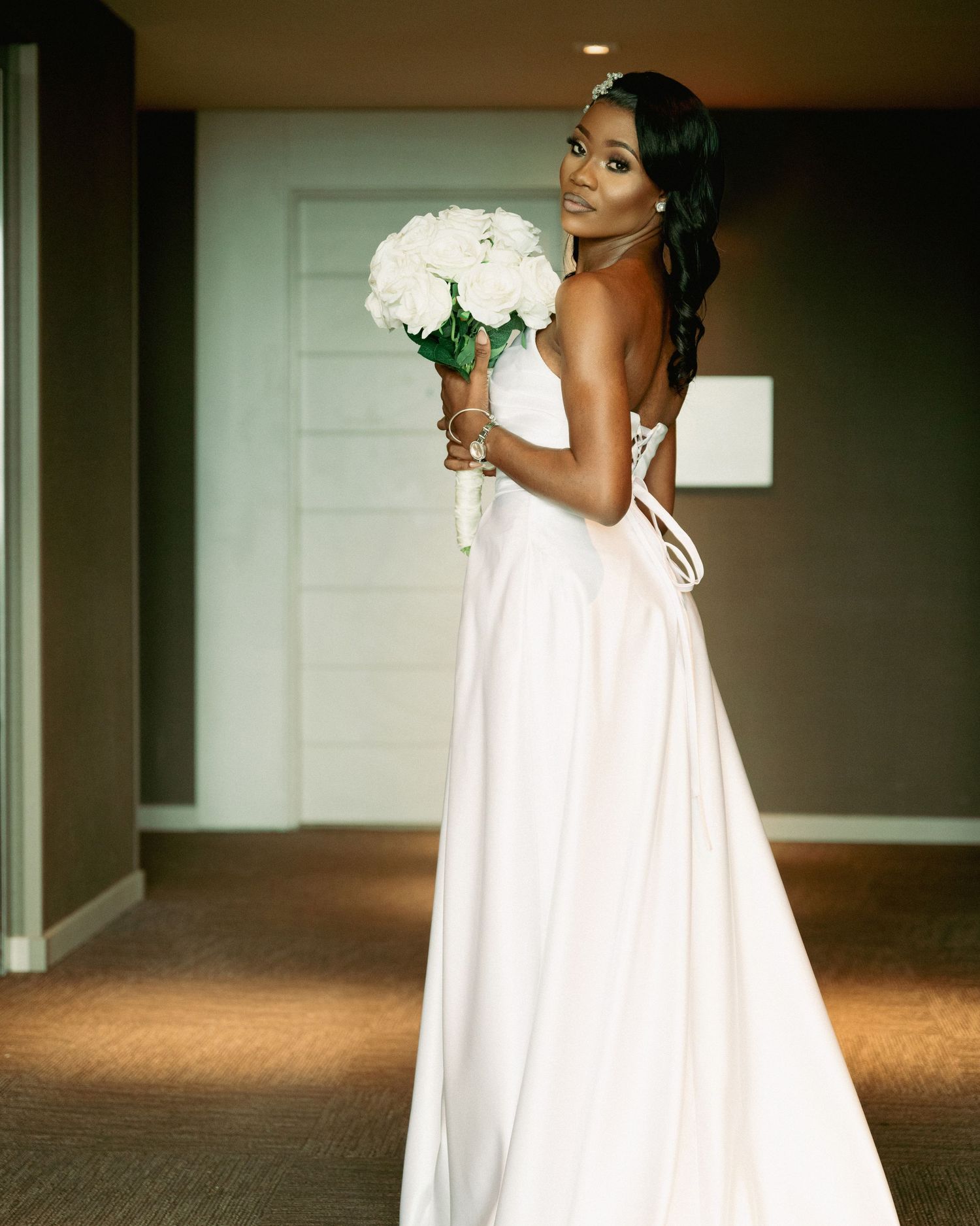 Bride in flowing white gown walks gracefully through indoor venue hallway.