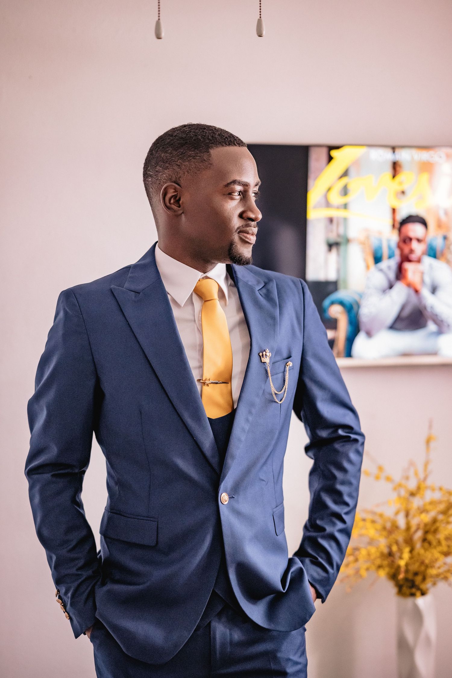 Groom in navy blue suit poses confidently against pink wall with yellow floral decor.