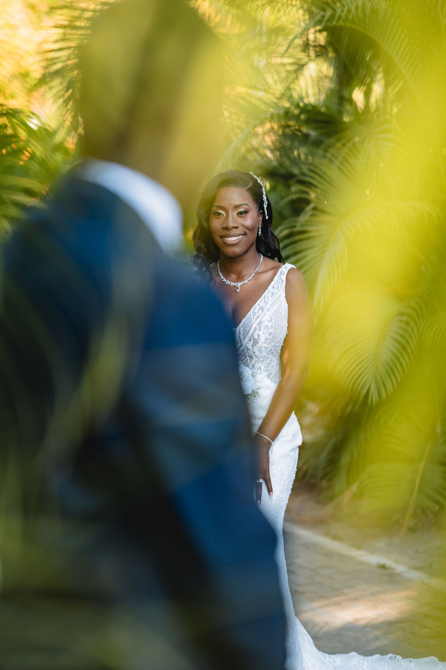 Wedding photography through palm fronds showing glimpses of a couple in white and navy attire.