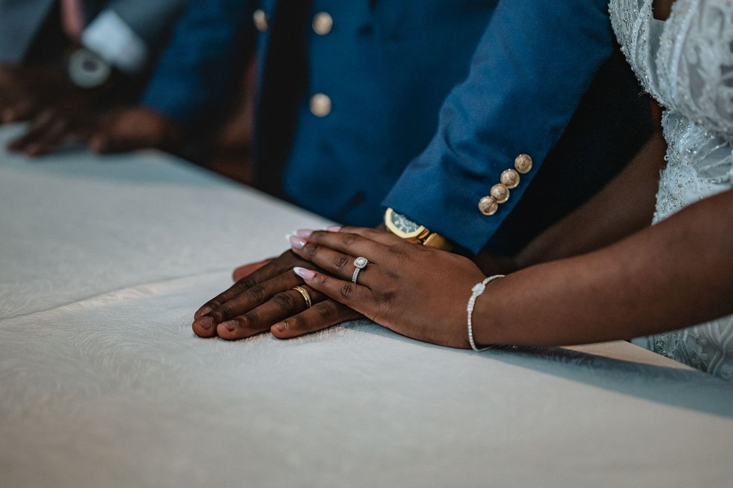 Close-up of Black couple's hands on white table, bride wearing diamond ring and gold bracelet, groom in blue suit.