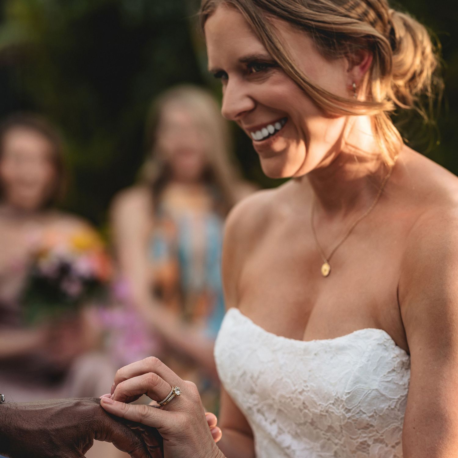 Smiling bride in strapless lace gown having ring placed on finger during outdoor wedding ceremony.