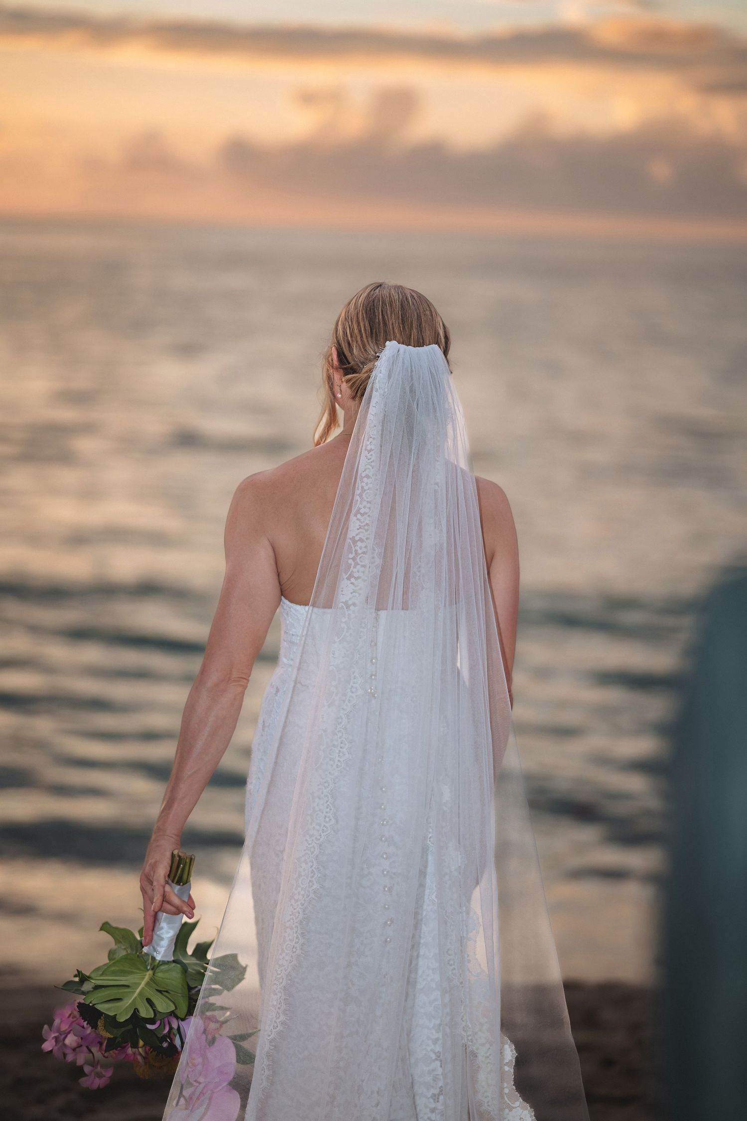 Bride in white lace gown and veil holds bouquet, gazing at ocean sunset from behind.