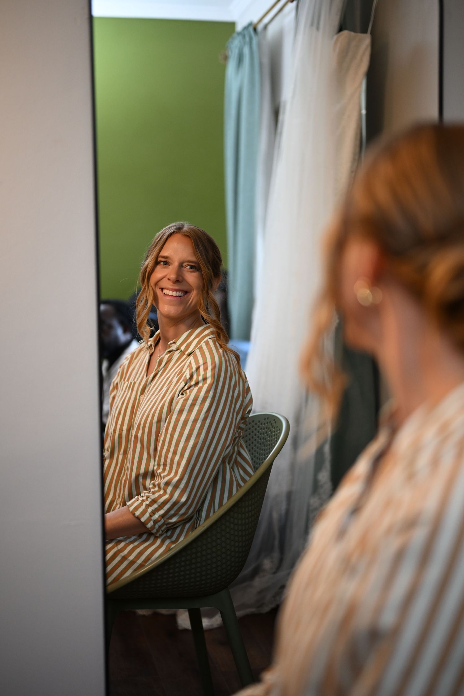 Smiling woman in striped shirt sitting in chair reflected in mirror inside a fitting room with green wall.