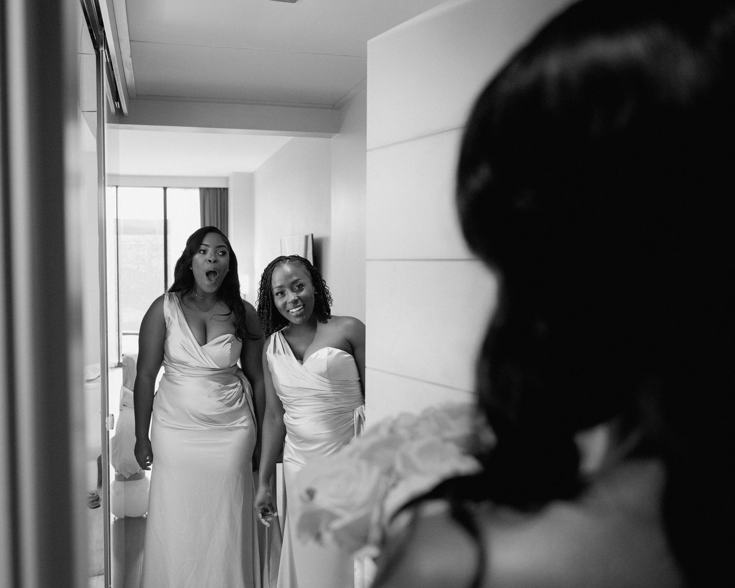 Black and white photo of two bridesmaids in white dresses reacting with joy upon seeing the bride.