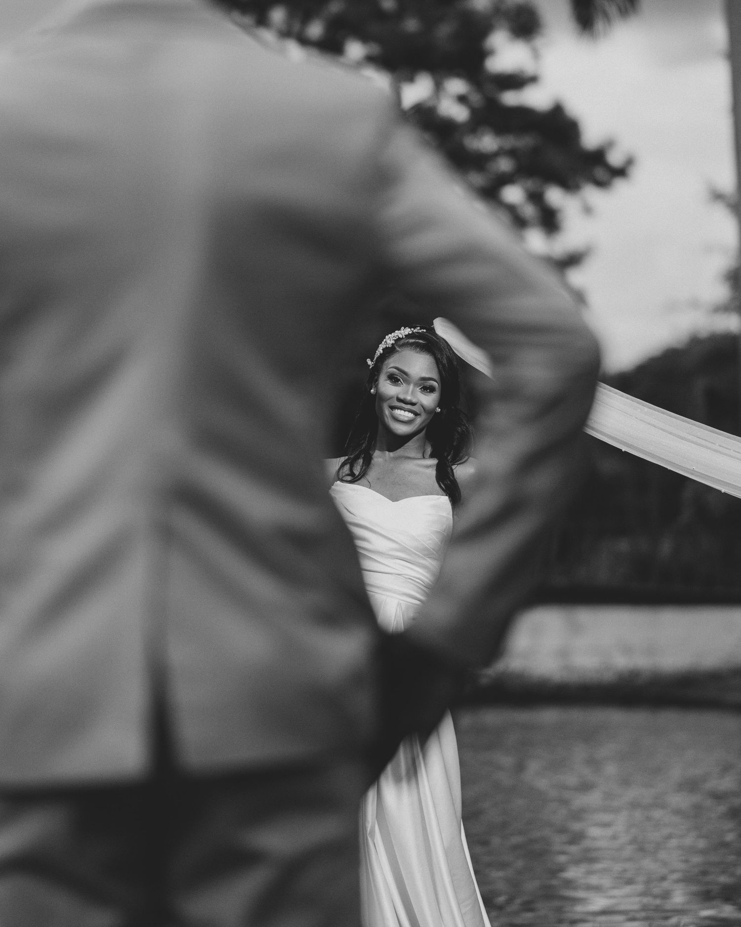 Black and white photo of smiling bride in wedding gown holding veil with groom in foreground.