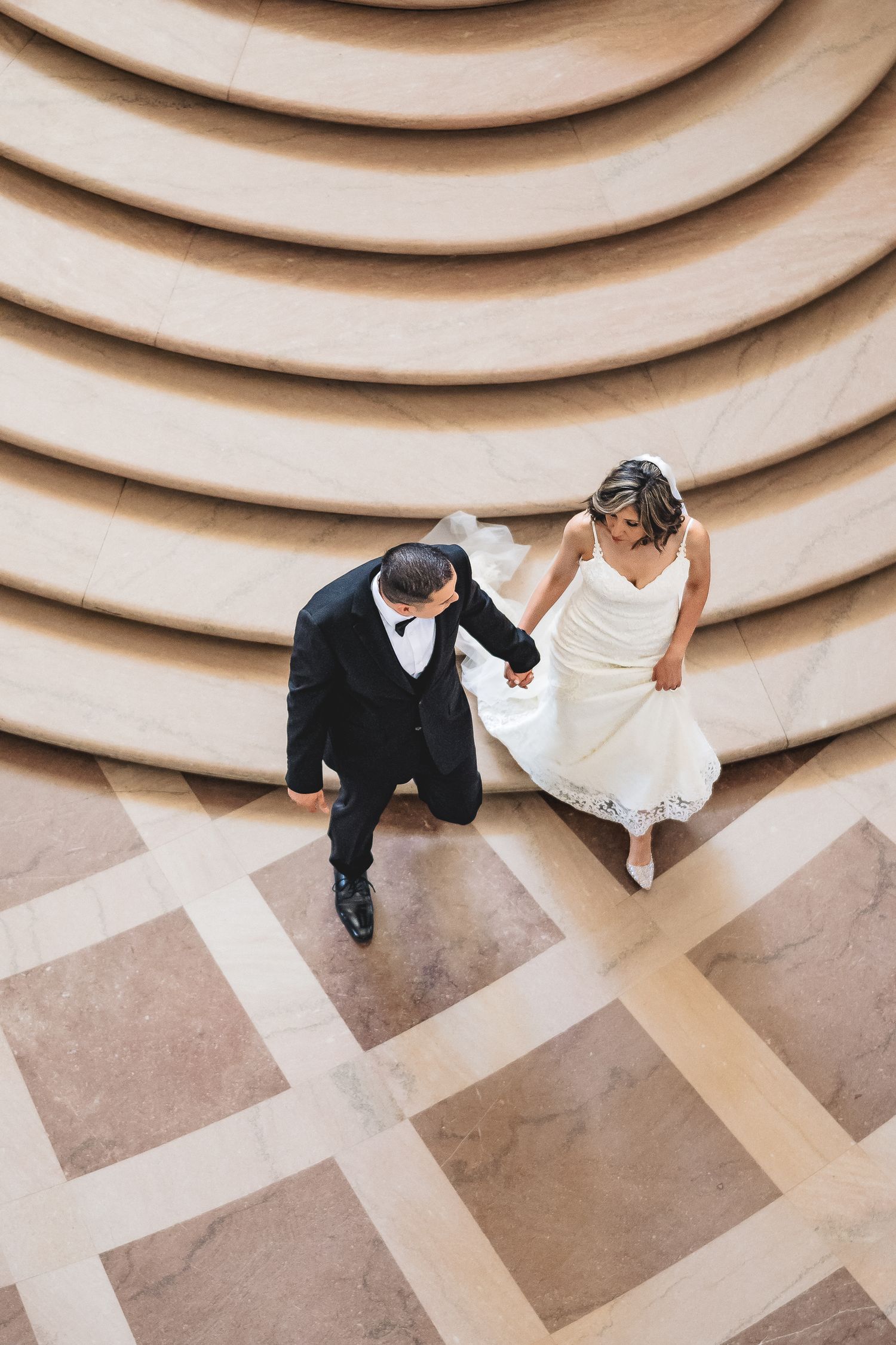 Aerial view of a bride and groom holding hands while walking across a marble floor near curved steps.