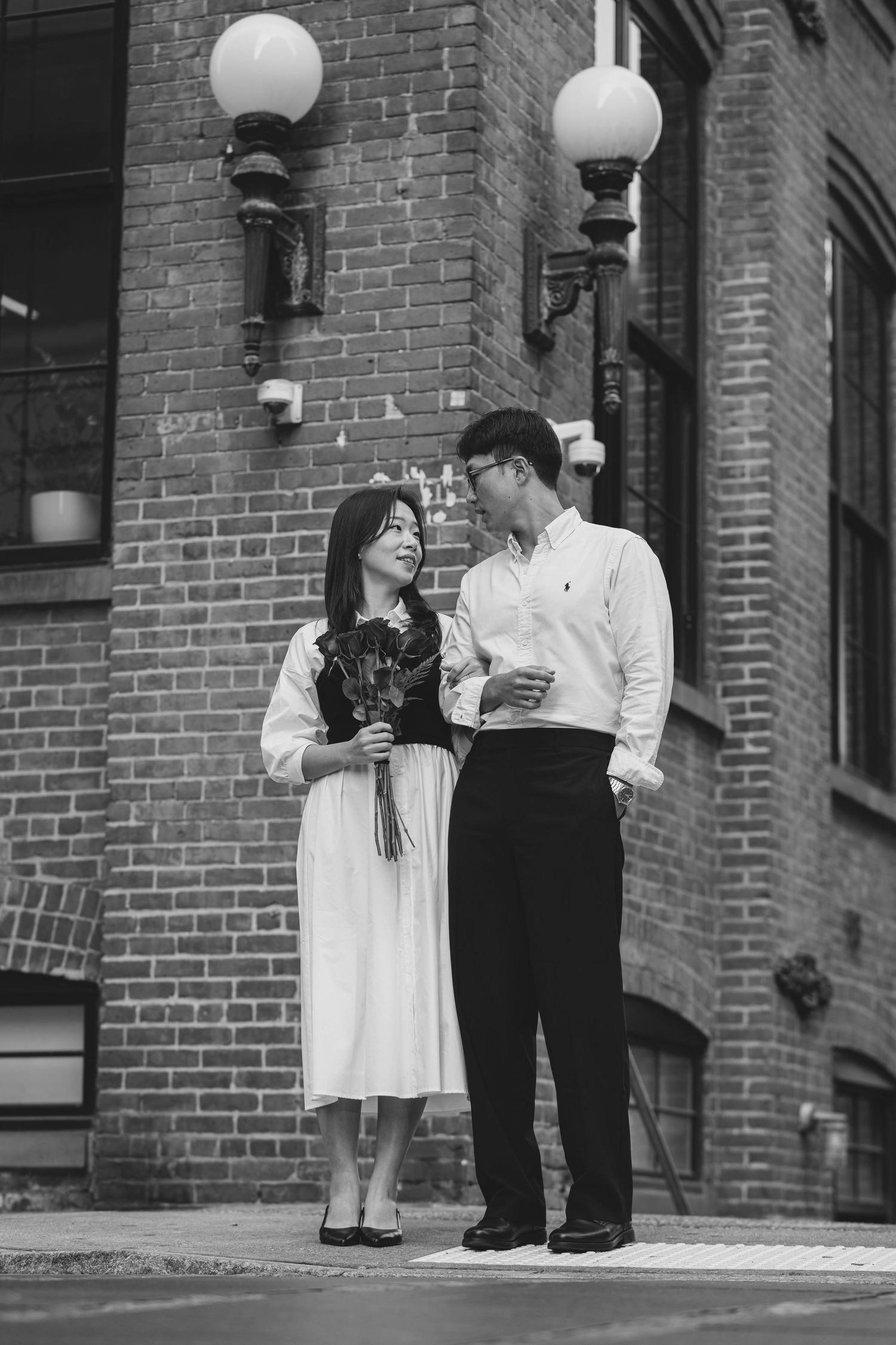Black and white photo of couple standing by brick building with vintage street lamp.