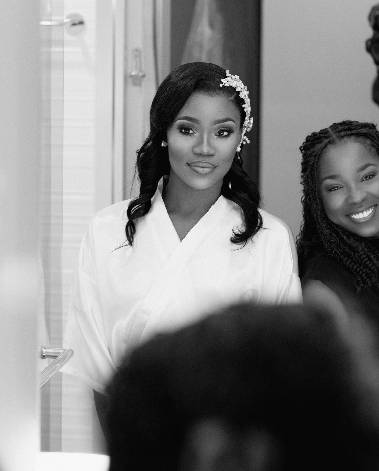 Black and white photo of a beautiful Black bride in white robe with floral headpiece getting ready with friends.