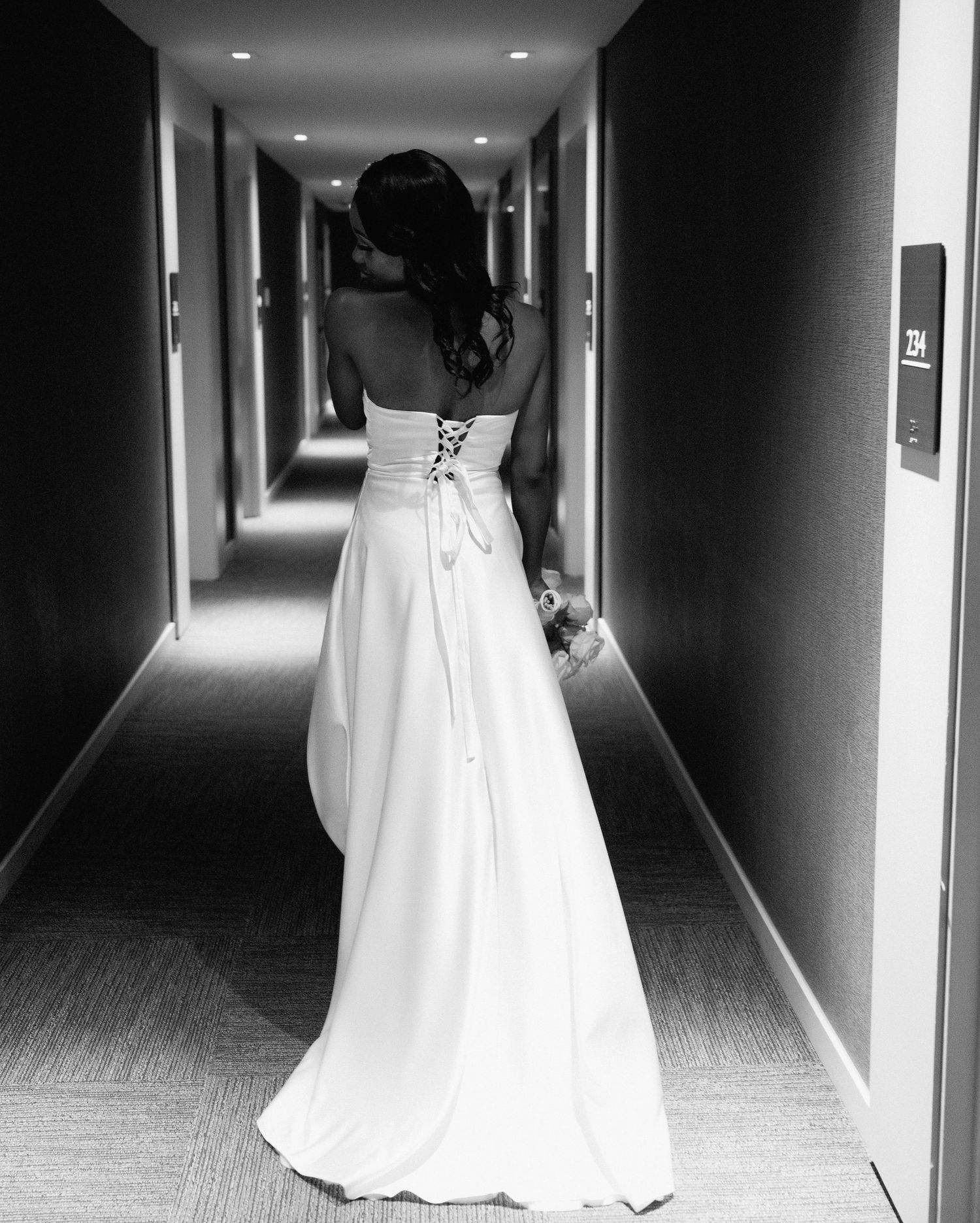 Black and white photo of bride in lace-back white gown walking down hotel hallway with bouquet.