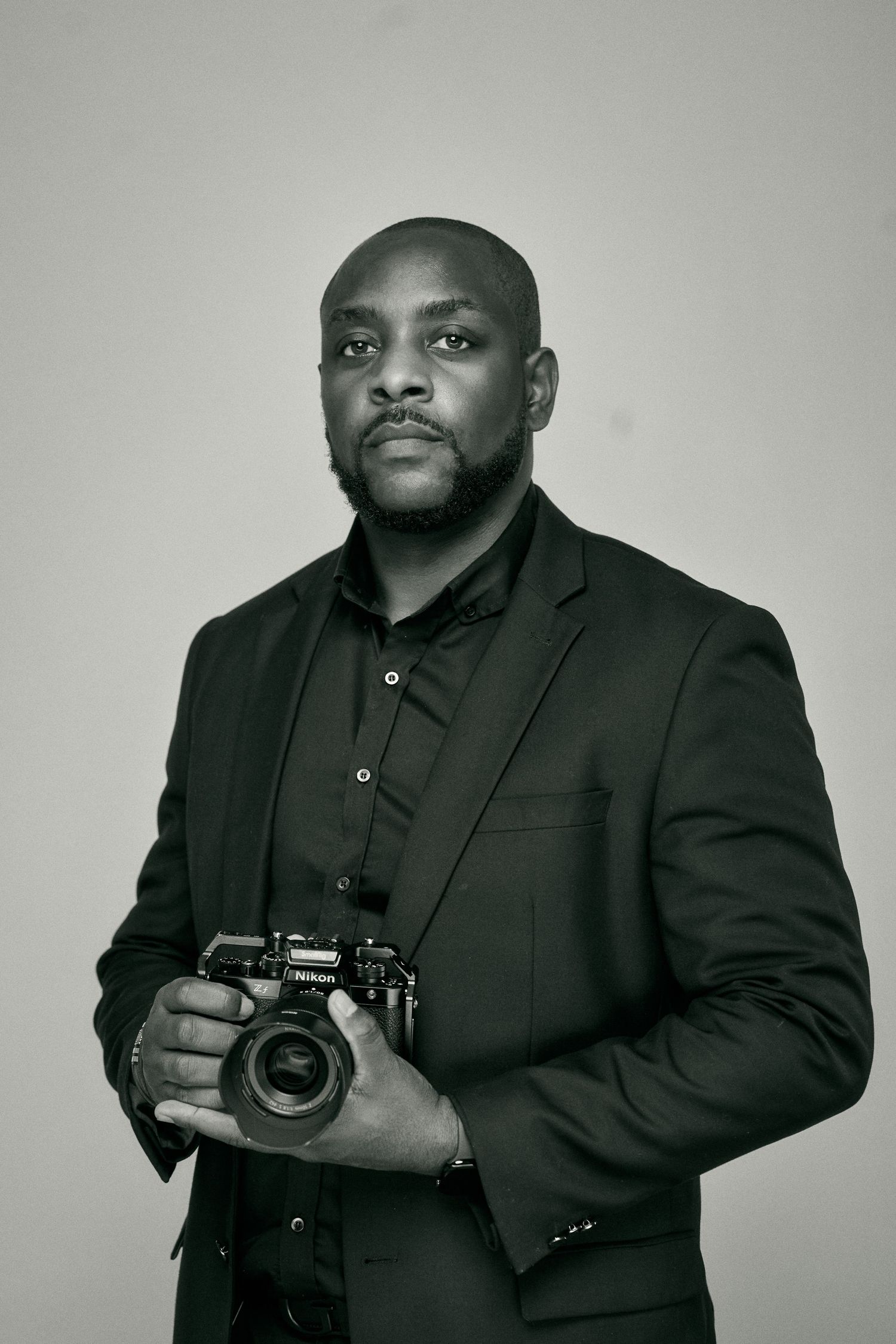 Professional photographer in black suit poses with camera against gray background.
