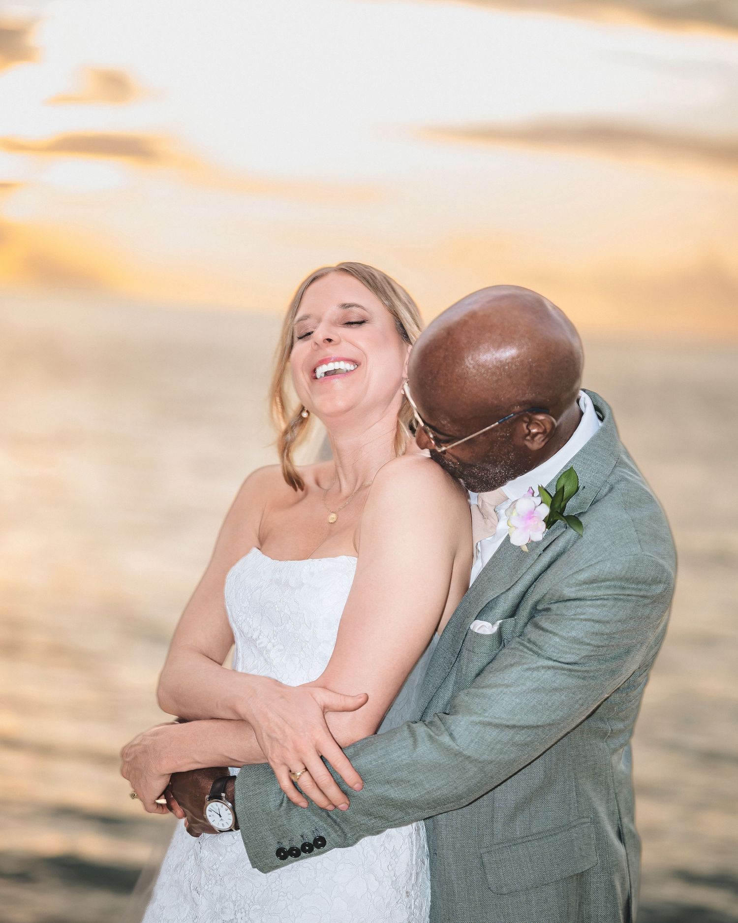 Bride and groom embrace at sunset on the beach, laughing joyfully in wedding attire with ocean backdrop.