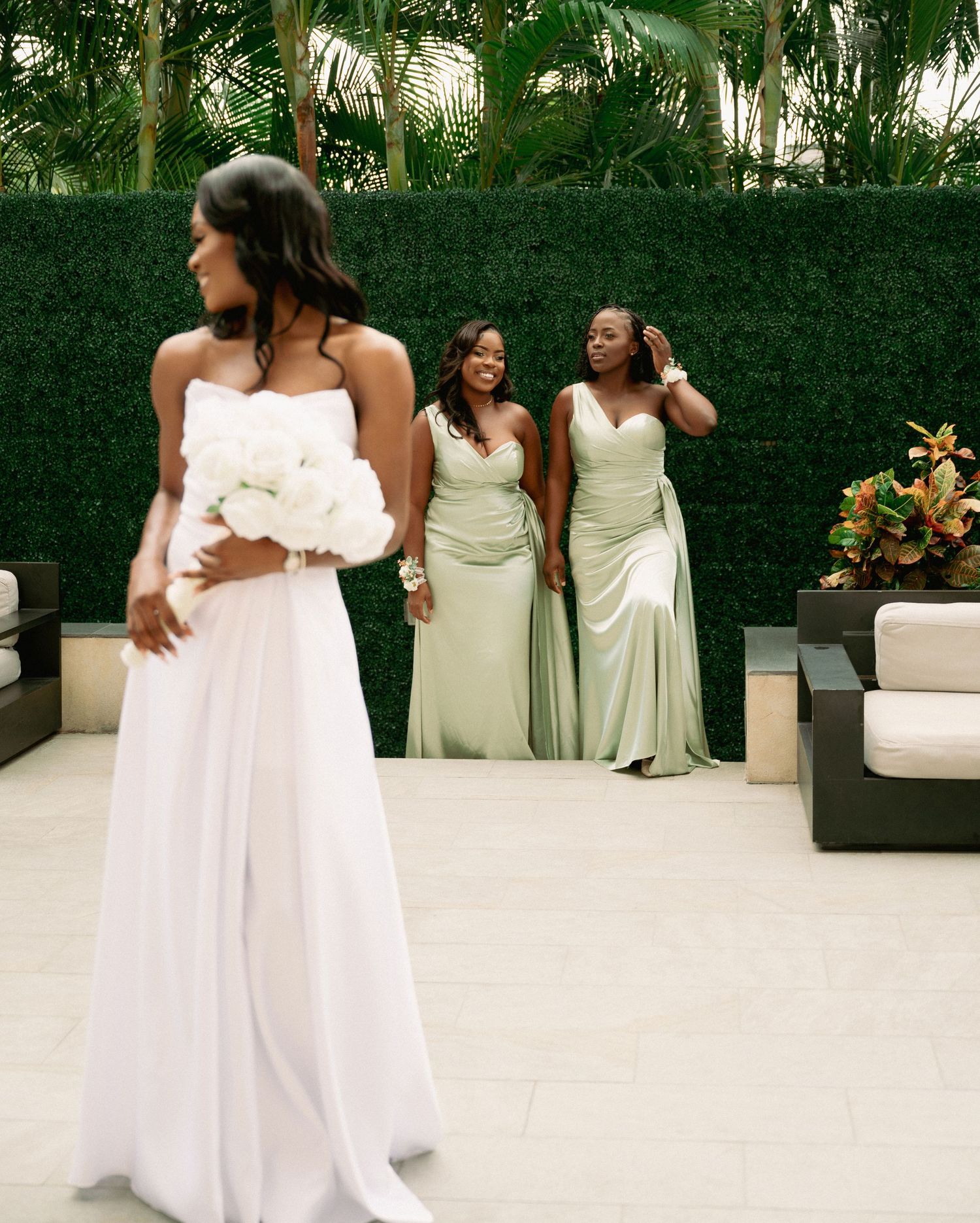 Bride holding white rose bouquet stands before two bridesmaids in sage green gowns against lush hedge backdrop.