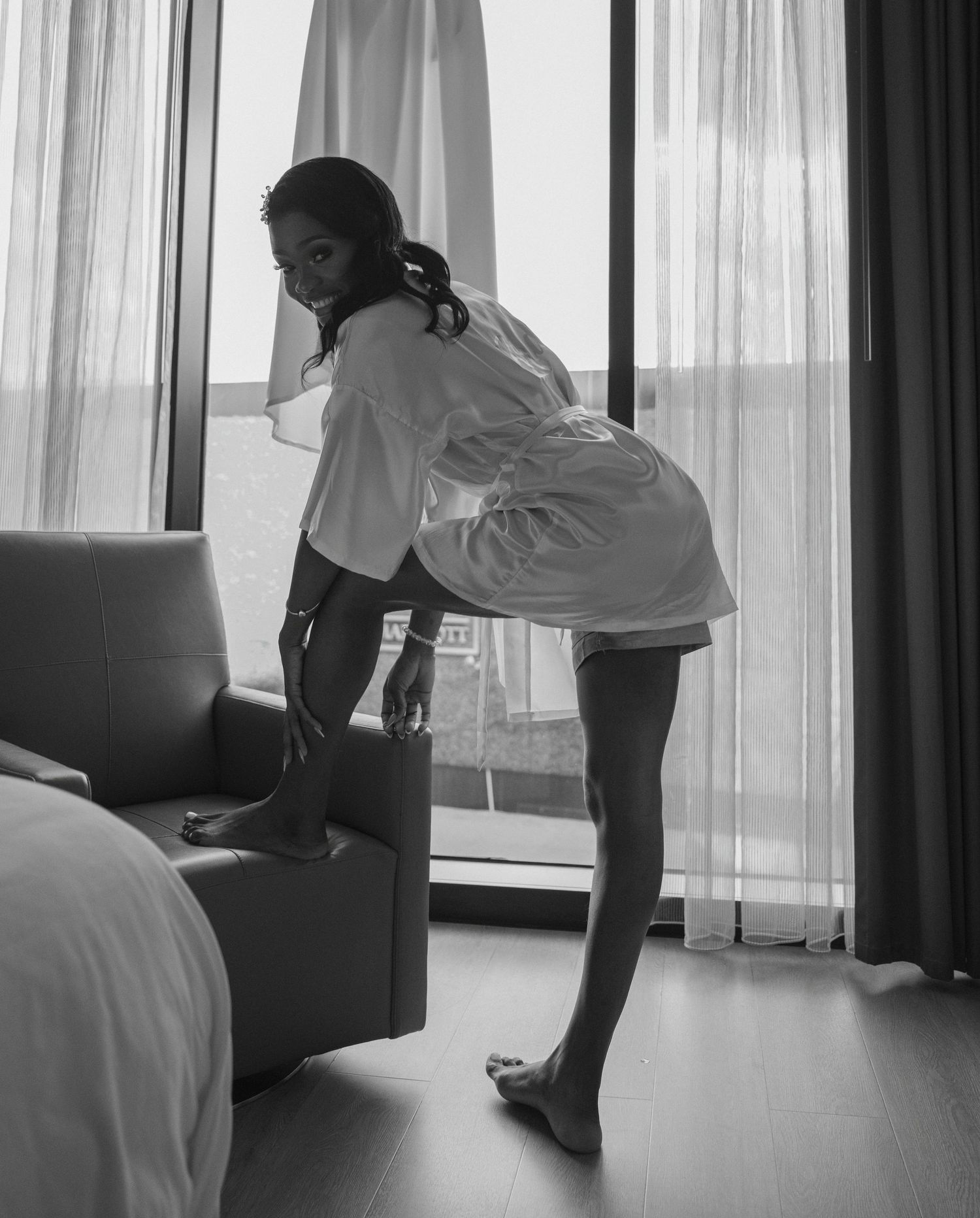 Black and white photo of a bride in a white robe preparing in a hotel room on her wedding day.