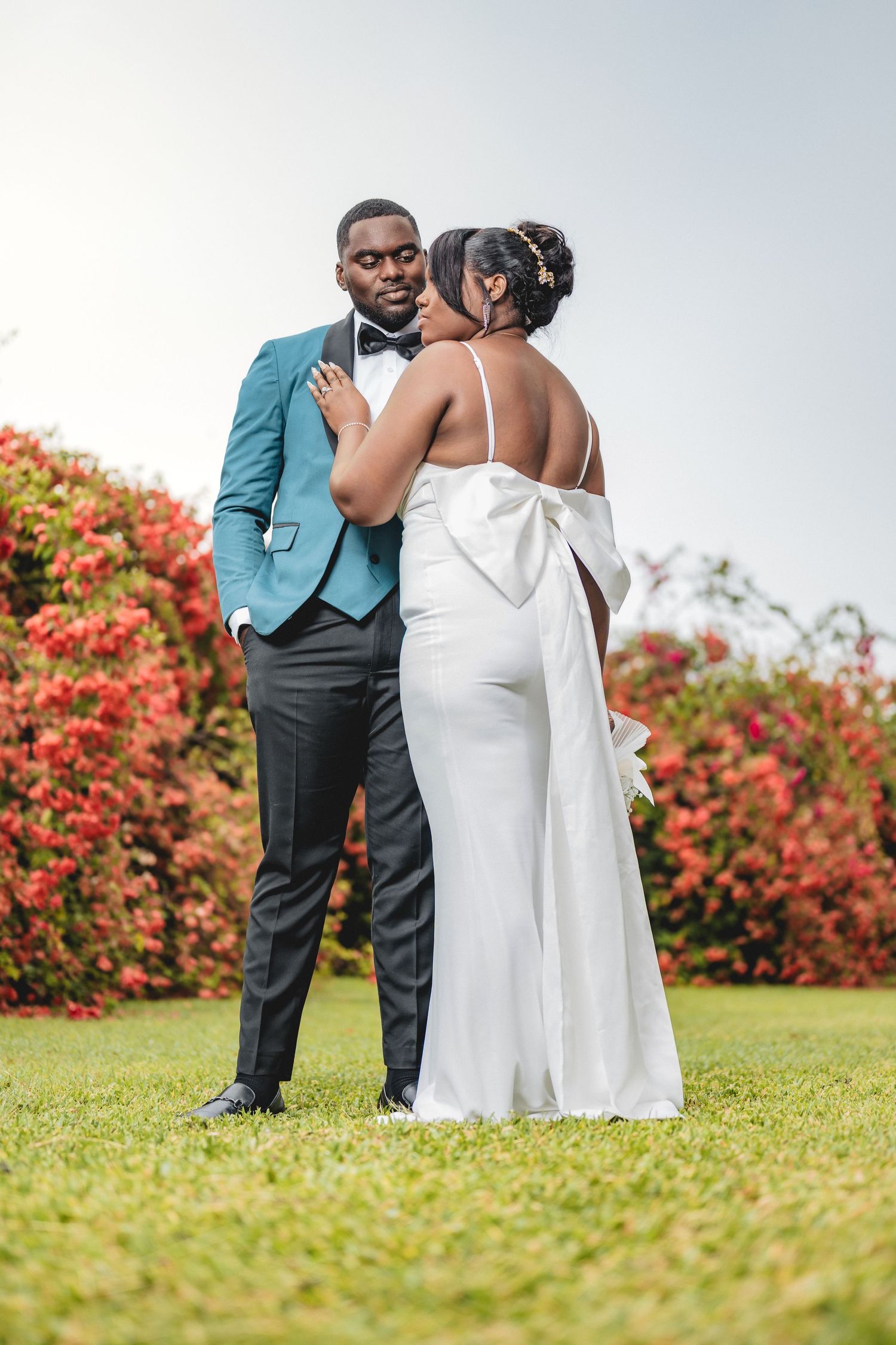 Elegant couple in wedding attire posing outdoors, groom in teal suit and bride in white backless gown with bow.