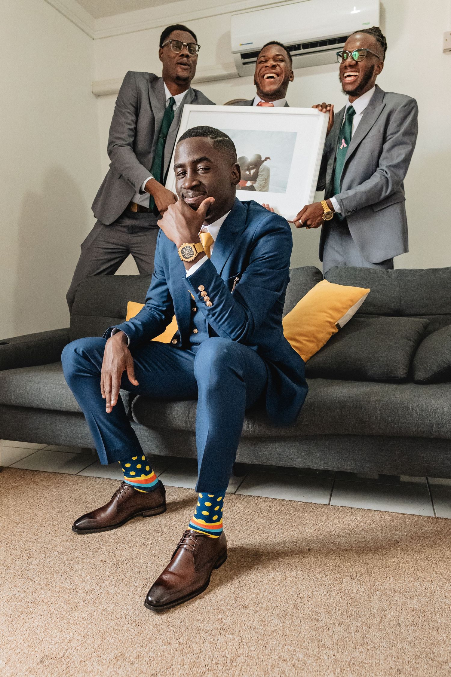 Groom in blue suit with colorful socks sits on sofa while groomsmen in gray suits hold framed artwork behind him.