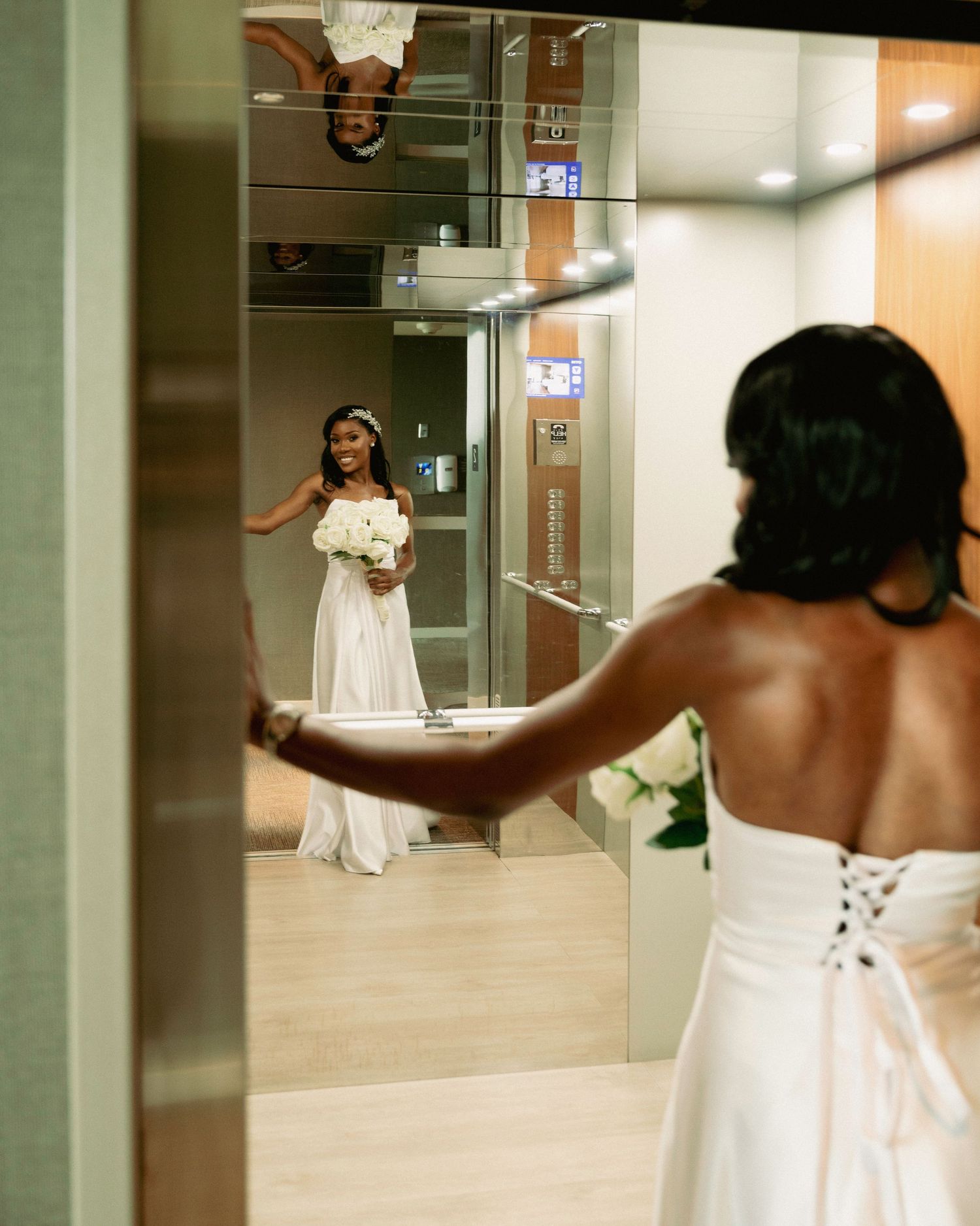 Bride in white gown holding bouquet reflected in elevator mirror on wedding day.