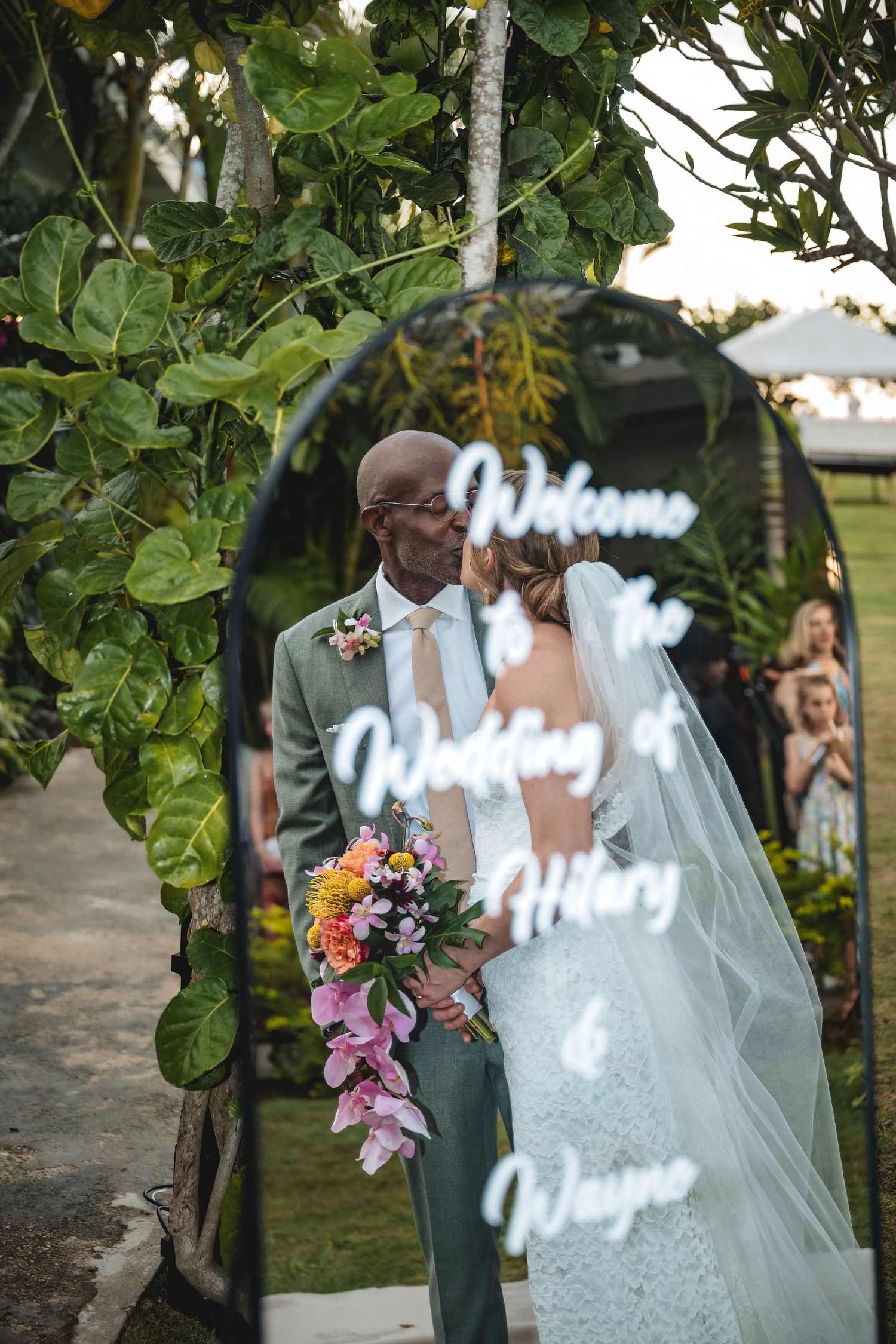 Interracial couple sharing a kiss at their outdoor wedding ceremony under a floral arch with a welcome sign reading Hilary and Wayne.