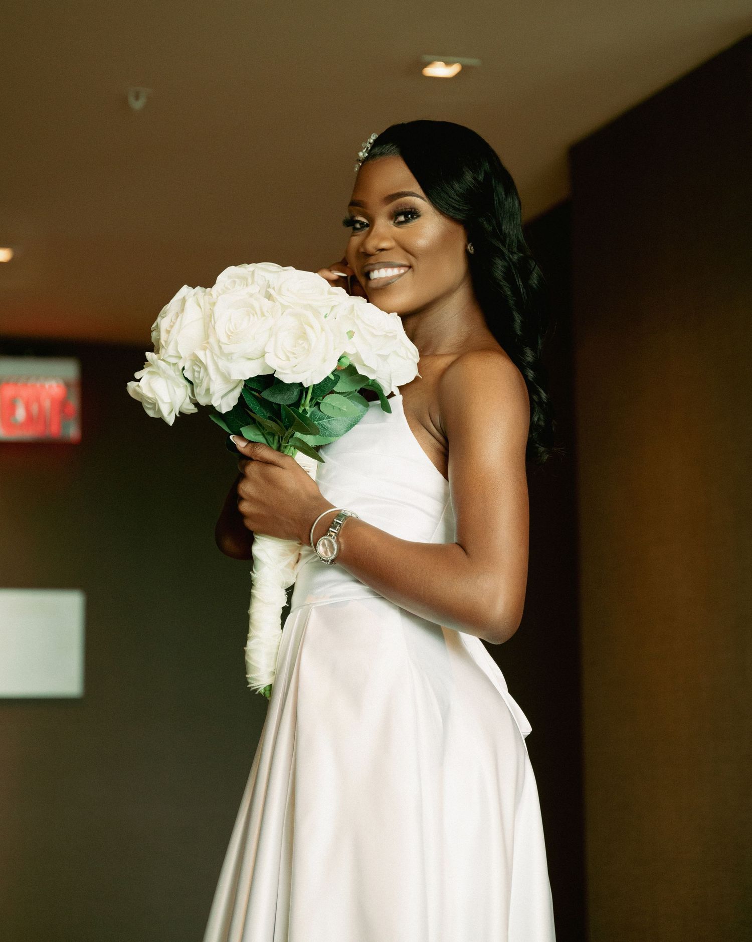 Smiling bride in white satin dress holds lush white rose bouquet, wearing a diamond bracelet indoors.