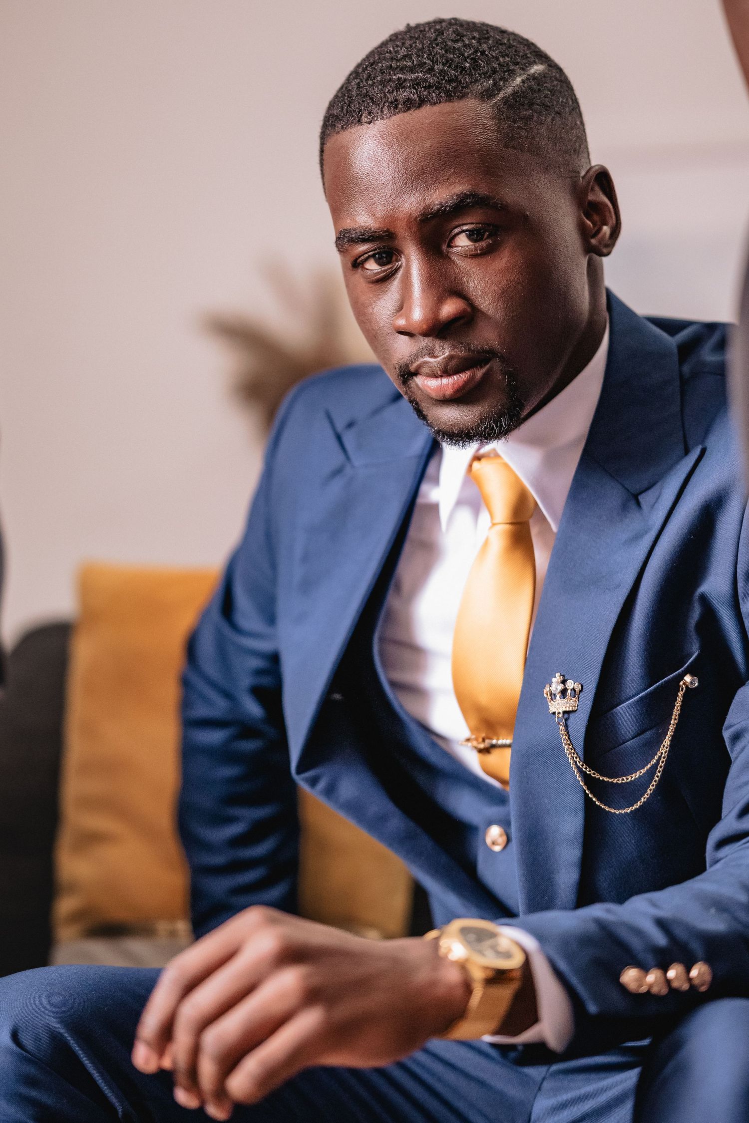 Elegant man in navy blue suit with gold tie and brooch checks his watch while seated.